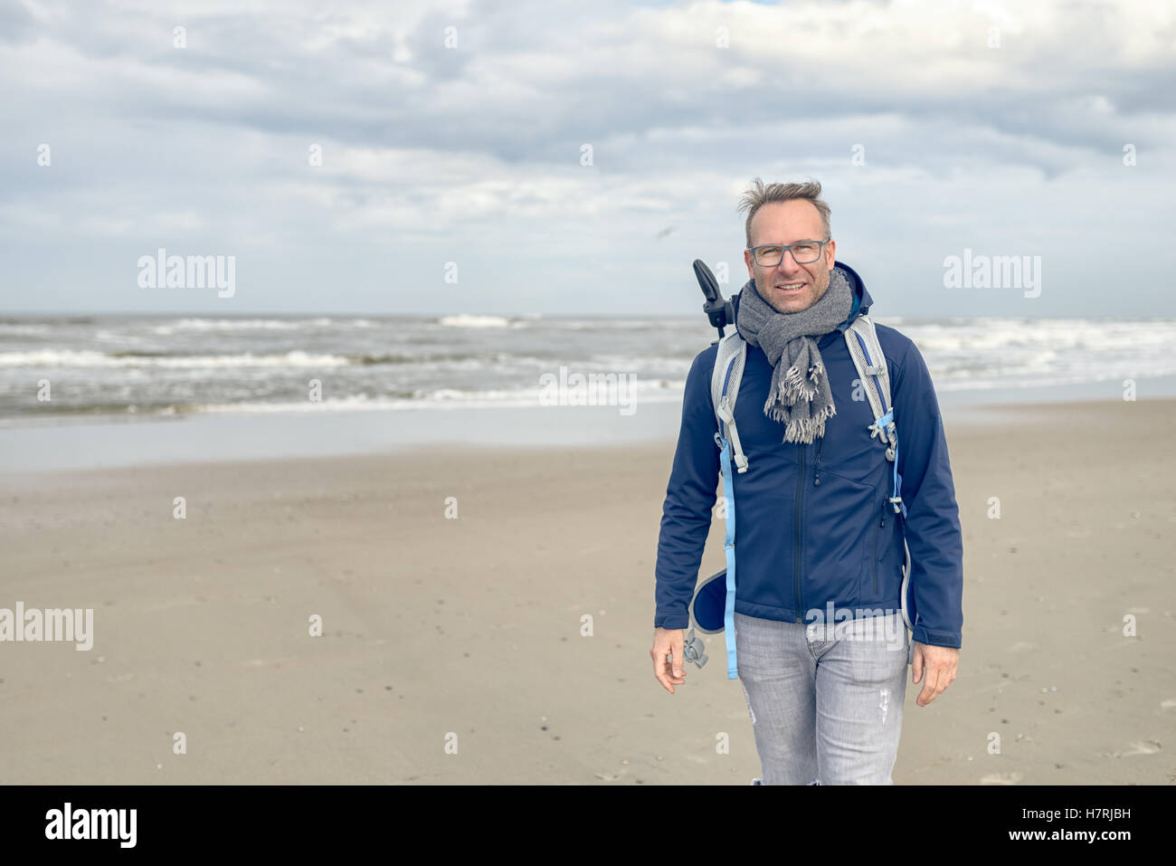 Im mittleren Alter Mann mit Brille, einen Rucksack und einen Schal zu Fuß entlang eines kalten blustery Strandes an einem regnerischen trüben Herbst oder winter Stockfoto