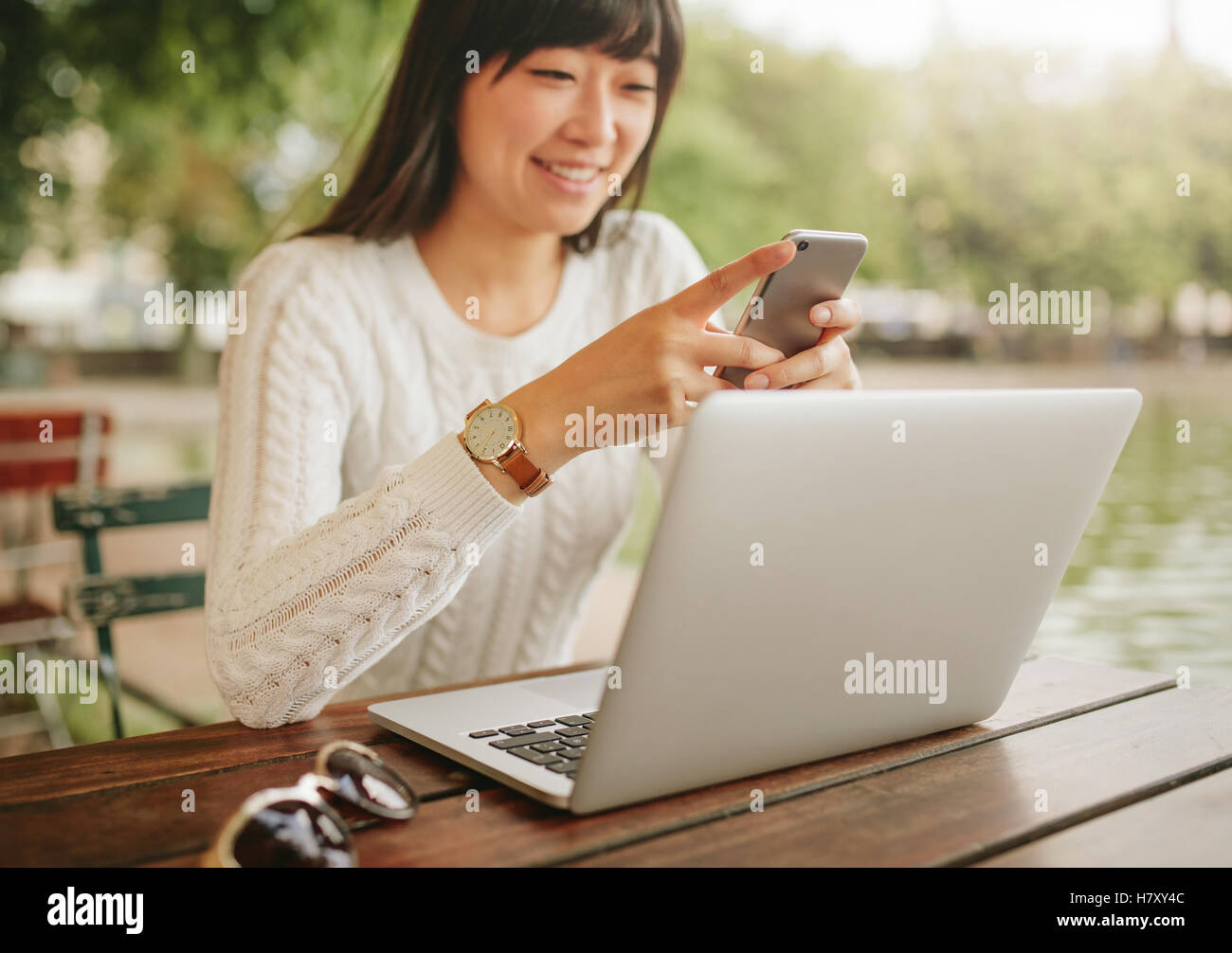 Schuss von glücklich Asiatin mit Smartphone auf Outdoor-Coffee-Shop. Frau mit Laptop im Freien mit Mobil am Café-Tisch sitzen Stockfoto