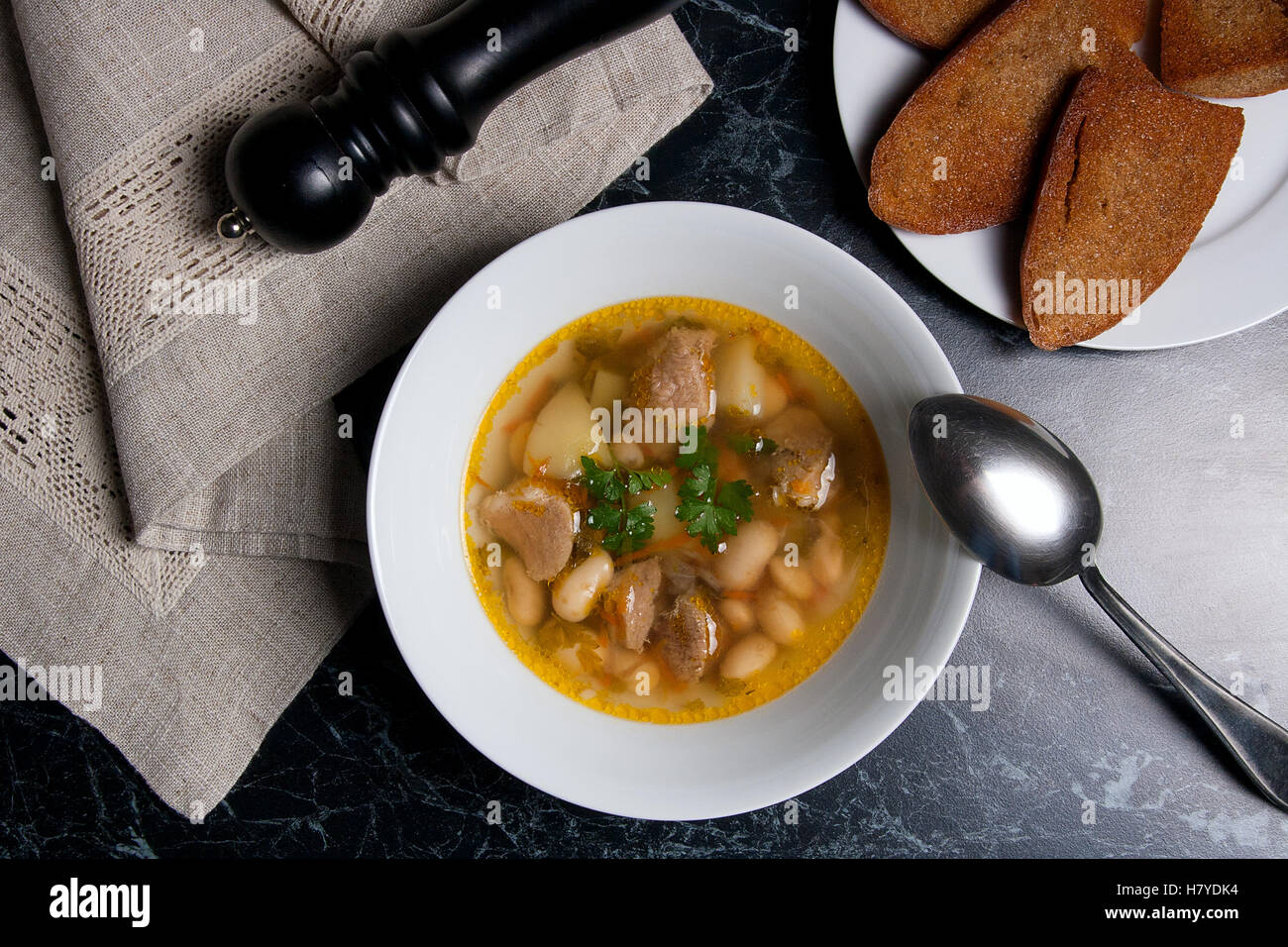 Bohnensuppe in weiße Schüssel mit Metalllöffel auf einem schwarzen Stein Hintergrund. Mehrere Popups auf weißen Teller und schwarze Mühle für Pfeffer Stockfoto