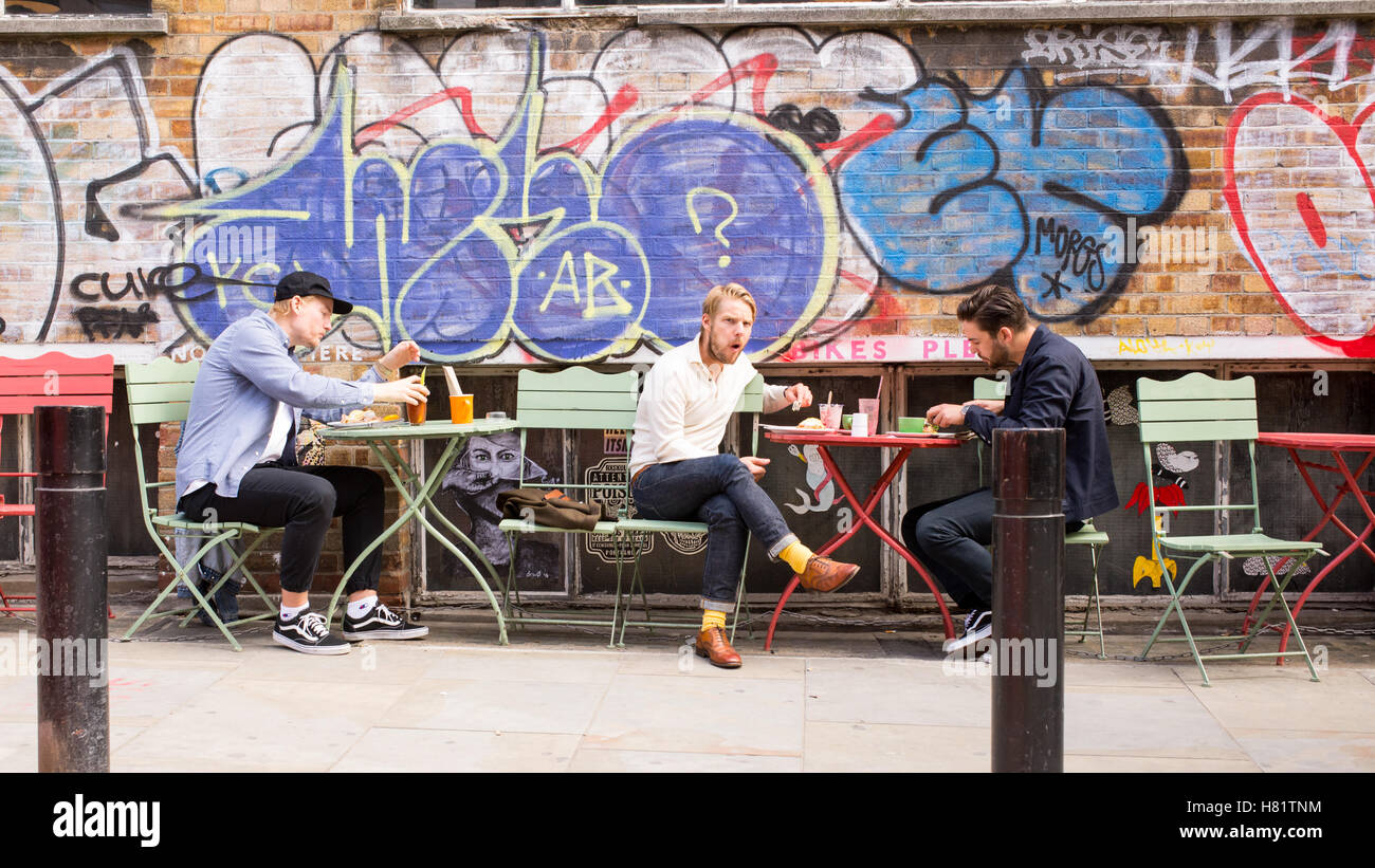 Junge Männer, die einen Brunch, draußen zu sitzen, vor einer Wand bedeckt Graffiti im kühlen Bereich der Shoreditch, East London Stockfoto