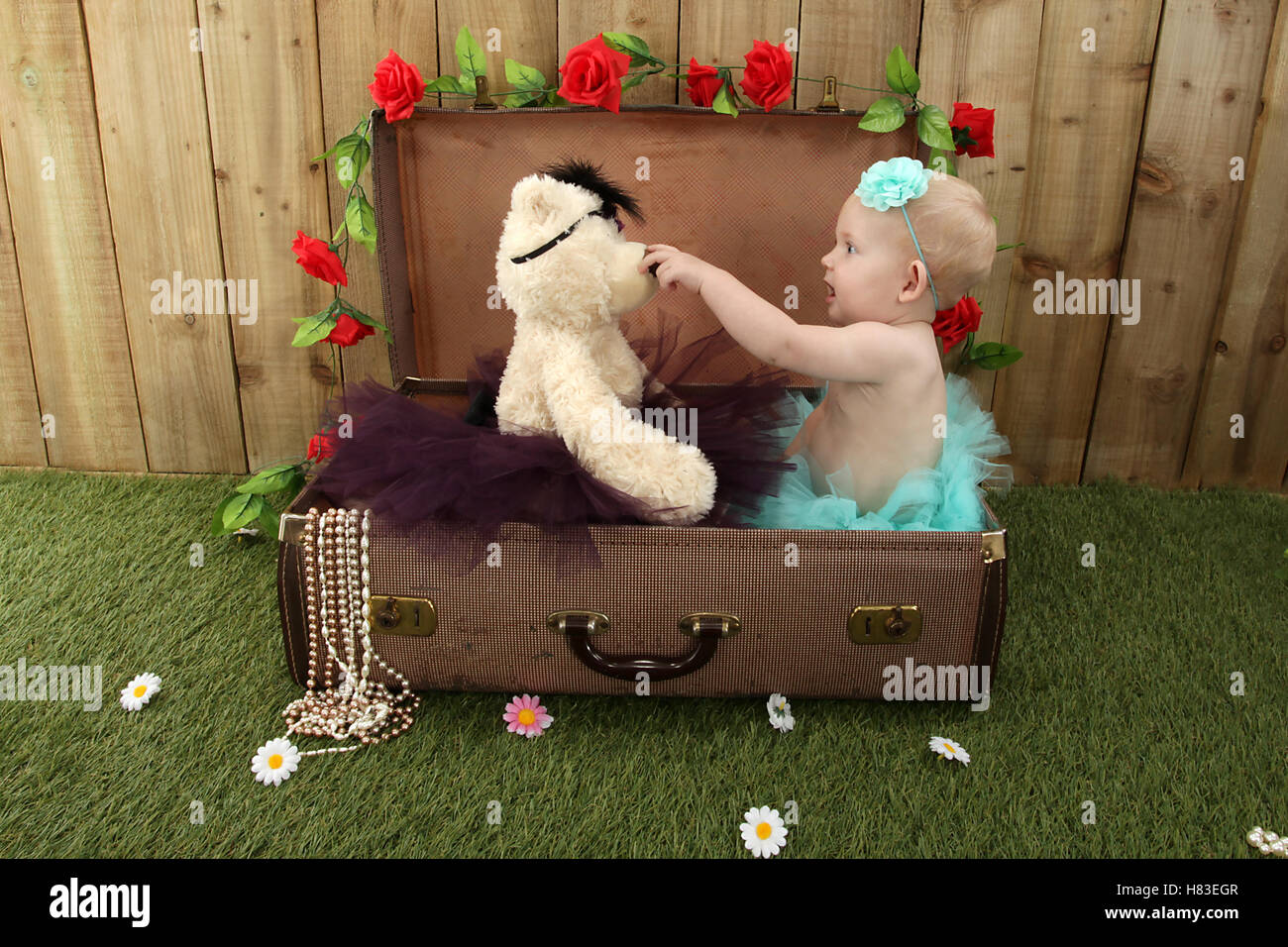 Babymädchen sitzen in einem Koffer im Garten mit Rasen und Blumen Stockfoto