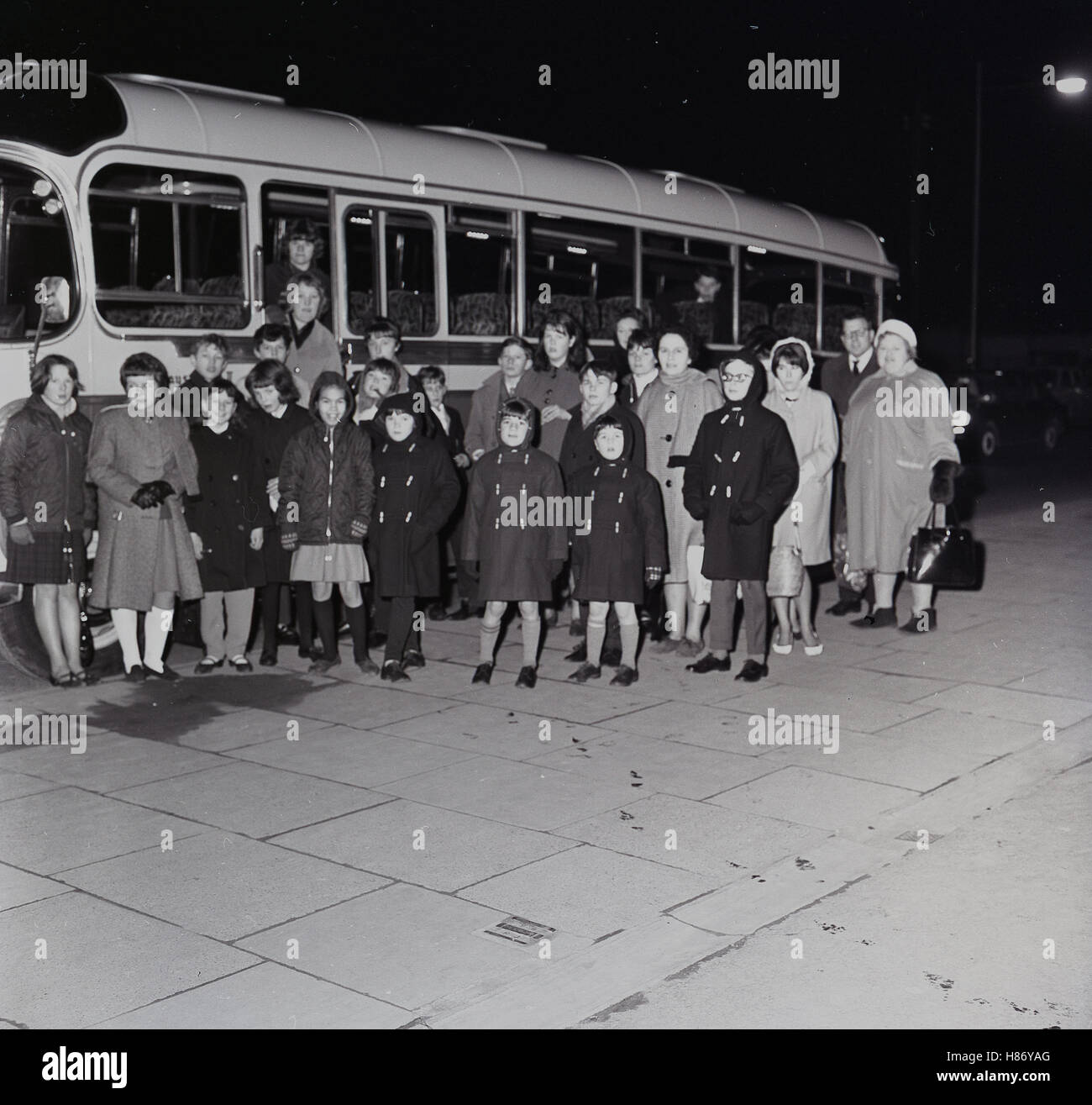 1965, aufgeregt historische, Abendzeit und eine Gruppe von kleinen Kindern im Wintermäntel mit begleitenden Erwachsenen-Line-up für ein Bild vor dem Einsteigen in einen Bus für einen Ausflug mit Übernachtung, England. Stockfoto