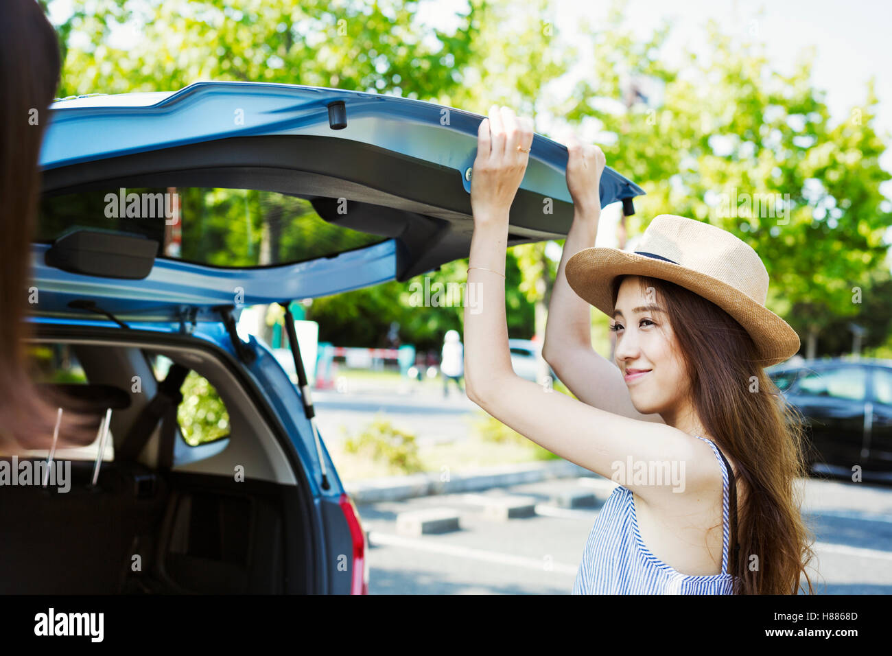 Lächelnde junge Frau mit langen braunen Haaren, Panama Hut trägt, schließen der Heckklappe eines Autos. Stockfoto