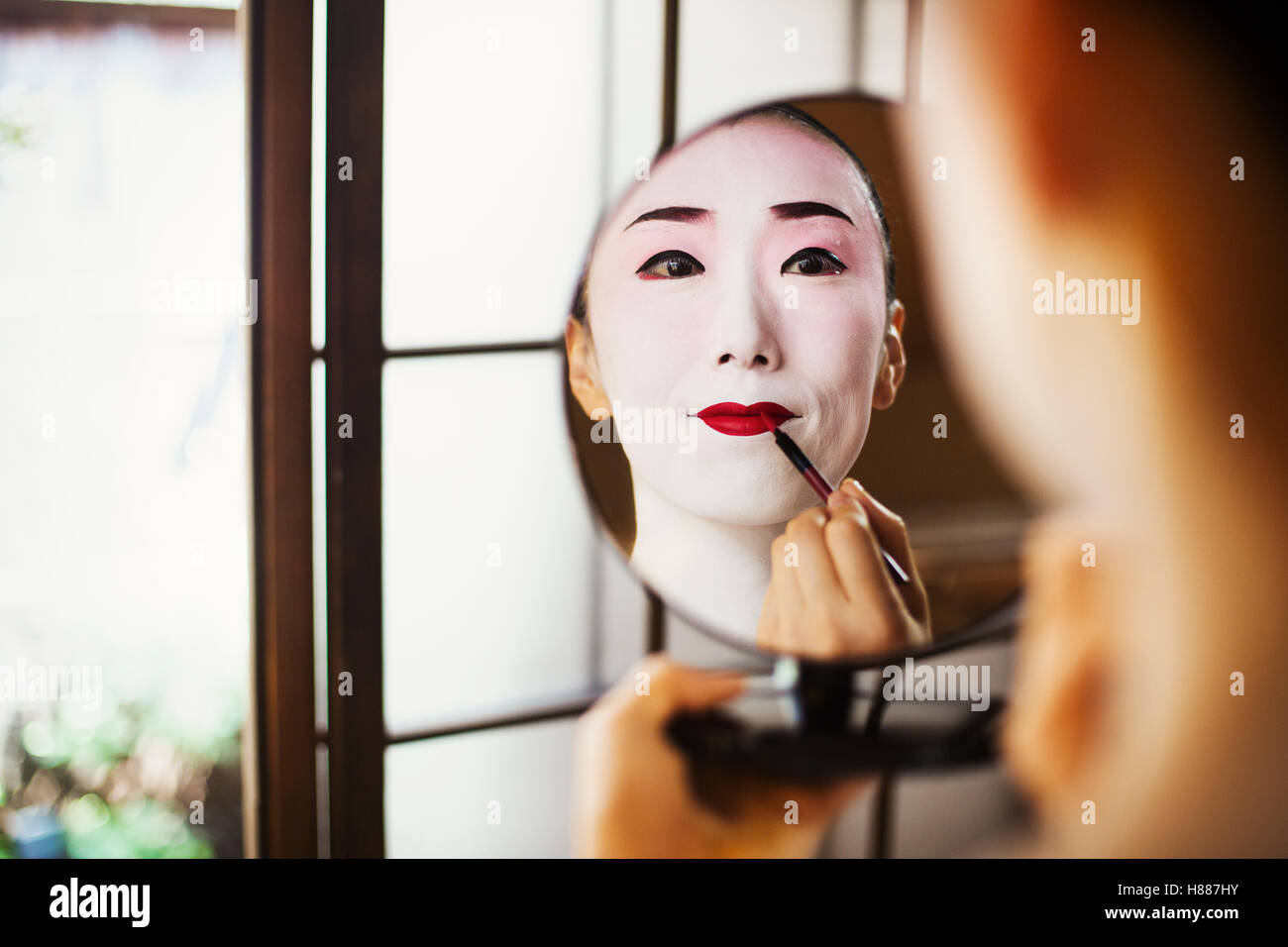 Geisha Frau mit traditionellen weißen Gesicht Make-up Anwendung knallroten Lippenstift mit einem Pinsel mit einem Spiegel. Stockfoto