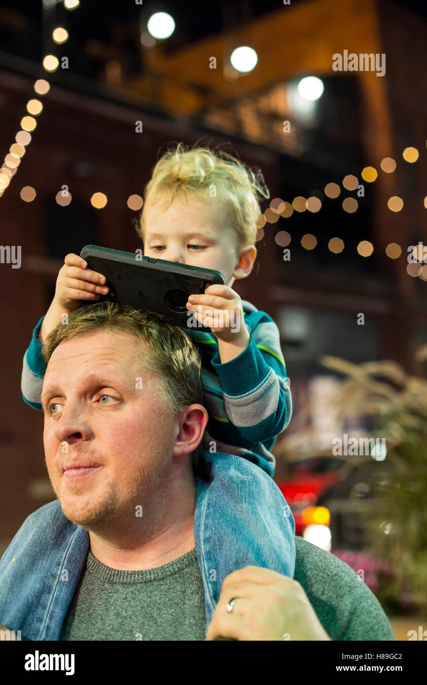 Detroit, Michigan - zwei-Jahr-alten Adam Hjermstad Jr. befasst sich mit einem Handy während der Fahrt auf den Schultern von seinem Vater. Stockfoto