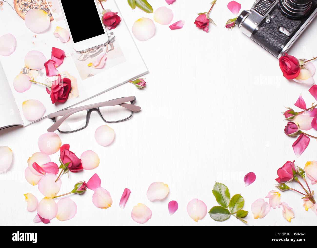 Die Zusammensetzung der roten Rosen, Blütenblätter, Magazin, Handy, Kamera auf einem weißen Hintergrund. Soft-Fokus. Ansicht von oben. Designer Stockfoto