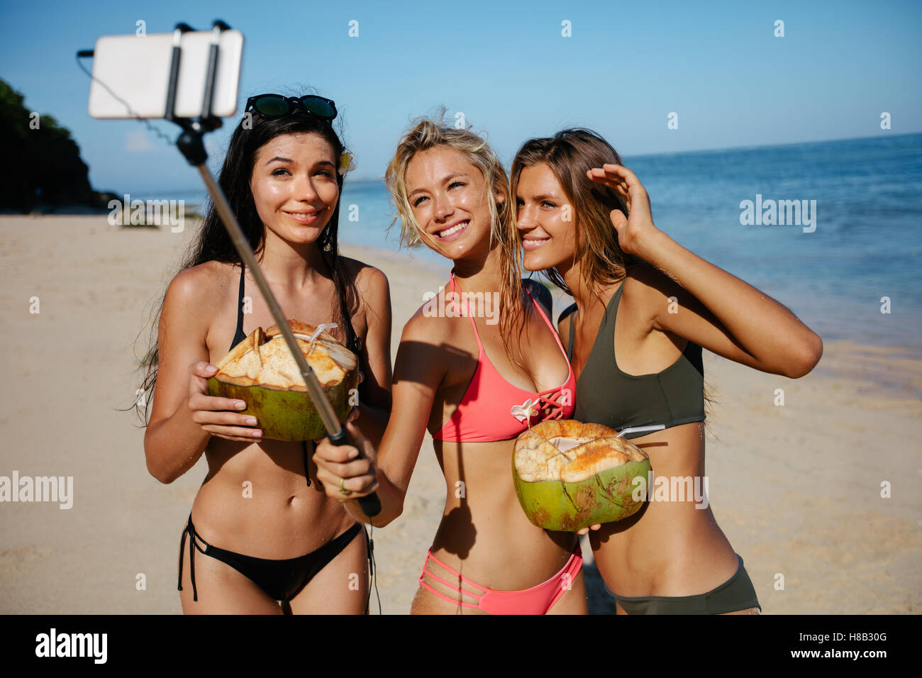 Drei junge Frauen im Badeanzug am Strand unter Selbstbildnis mit Smartphone auf Selfie Stick. Gruppe von Freundinnen holdin Stockfoto