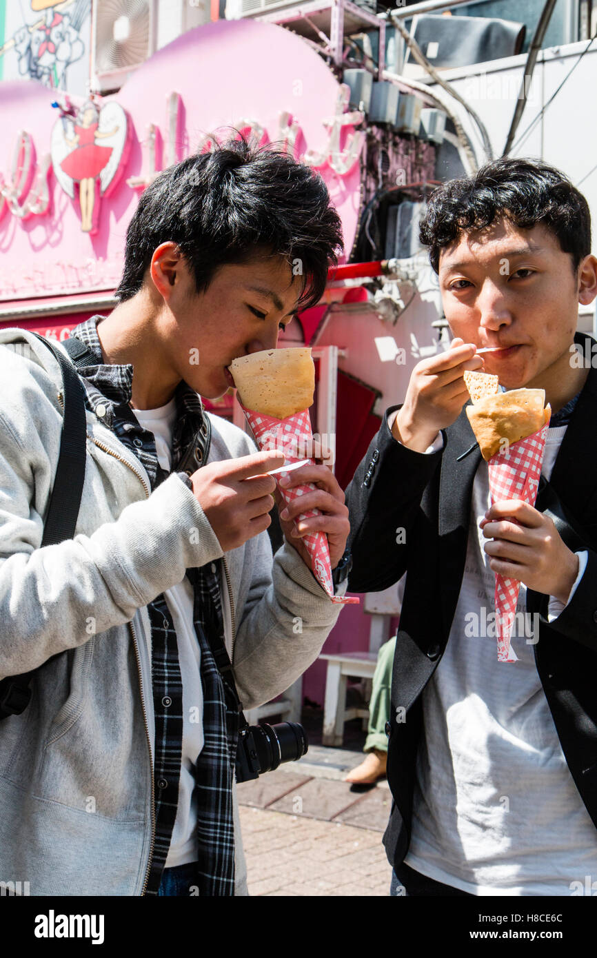 Japan, Tokio, Harajuku, Takeshita-dori. Zwei Teenager japanische Männer halten und essen Crepes in Kegel, auf Viewer. Angel's Herz Crêpe-Store. Stockfoto