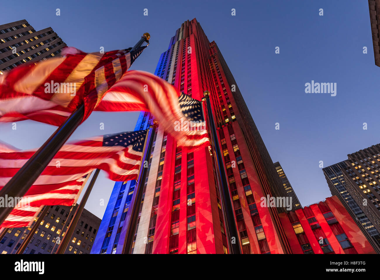 Rockefeller Center in der Dämmerung in weiß, rot und blau beleuchtet. Amerikanische Flaggen flattern im Wind. Midtown Manhattan, New York Stockfoto