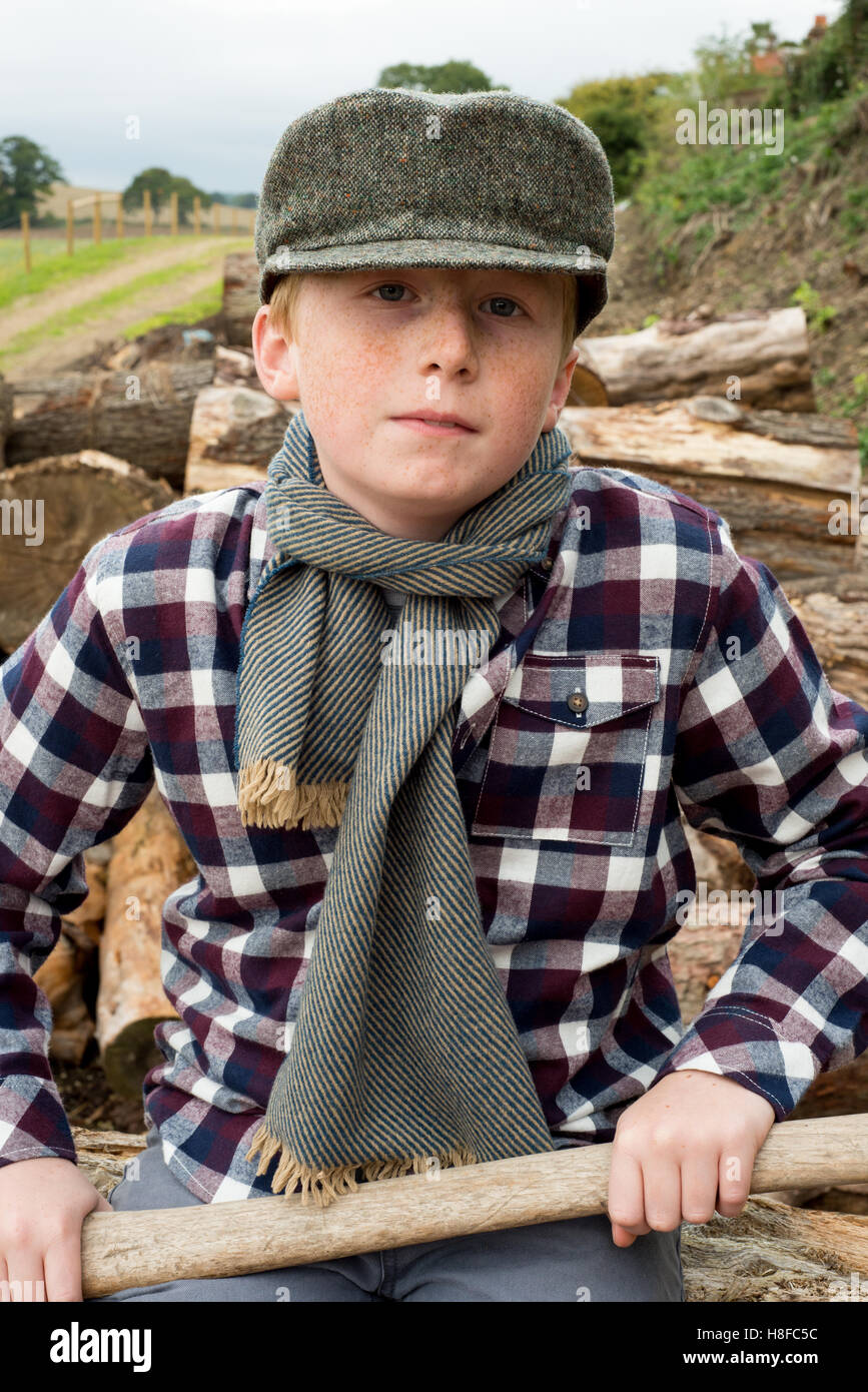 Junge im karierten Hemd, Schal und Mütze hält eine Axt vor einem Haufen Brennholz auf einem Bauernhof Stockfoto