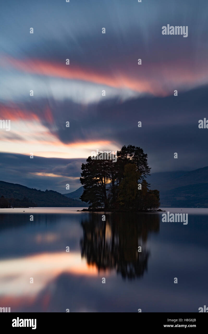 Loch Tay, Perthshire, Schottland, reflektiert das Licht von der untergehenden Sonne, wie es geht hinter den Bäumen auf einer der Inseln. Stockfoto