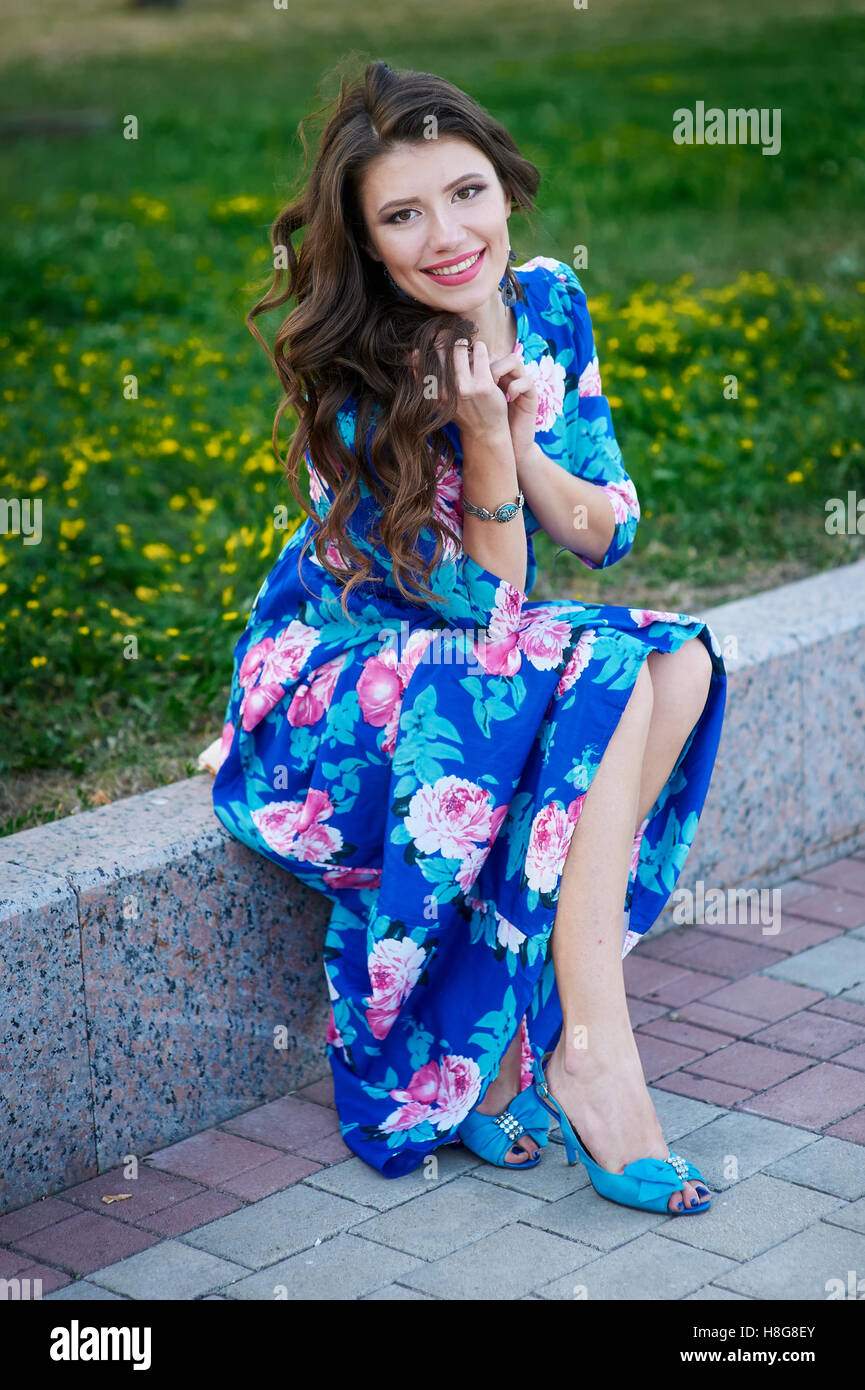 wunderschöne lächelnde Frau in einem blauen Kleid sitzt auf den Bordstein im Park Stockfoto