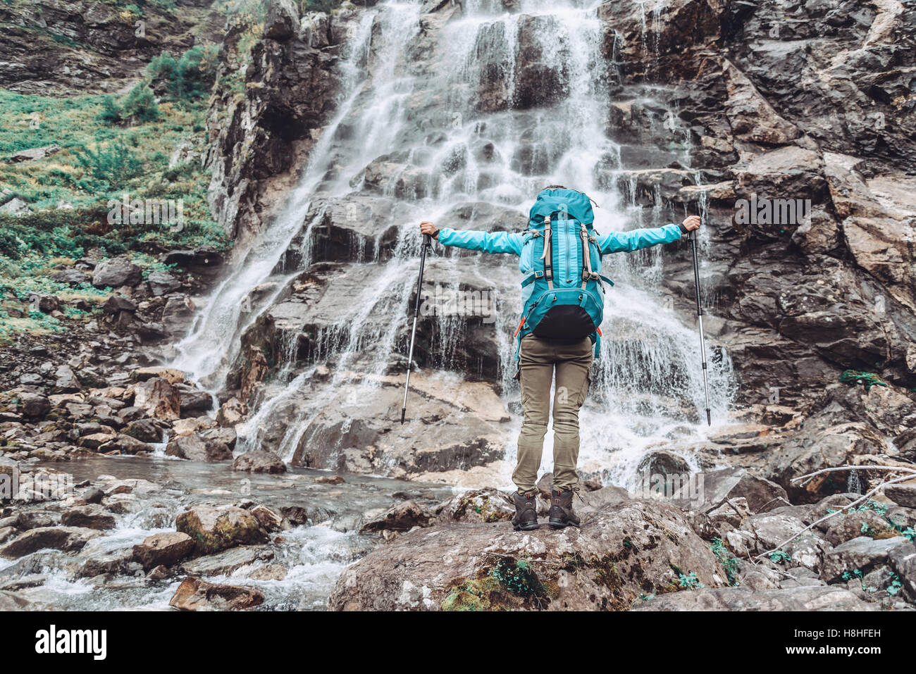 Glücklich Backpacker junge Frau auf Stein mit erhobenen Armen vor Wasserfall Stockfoto