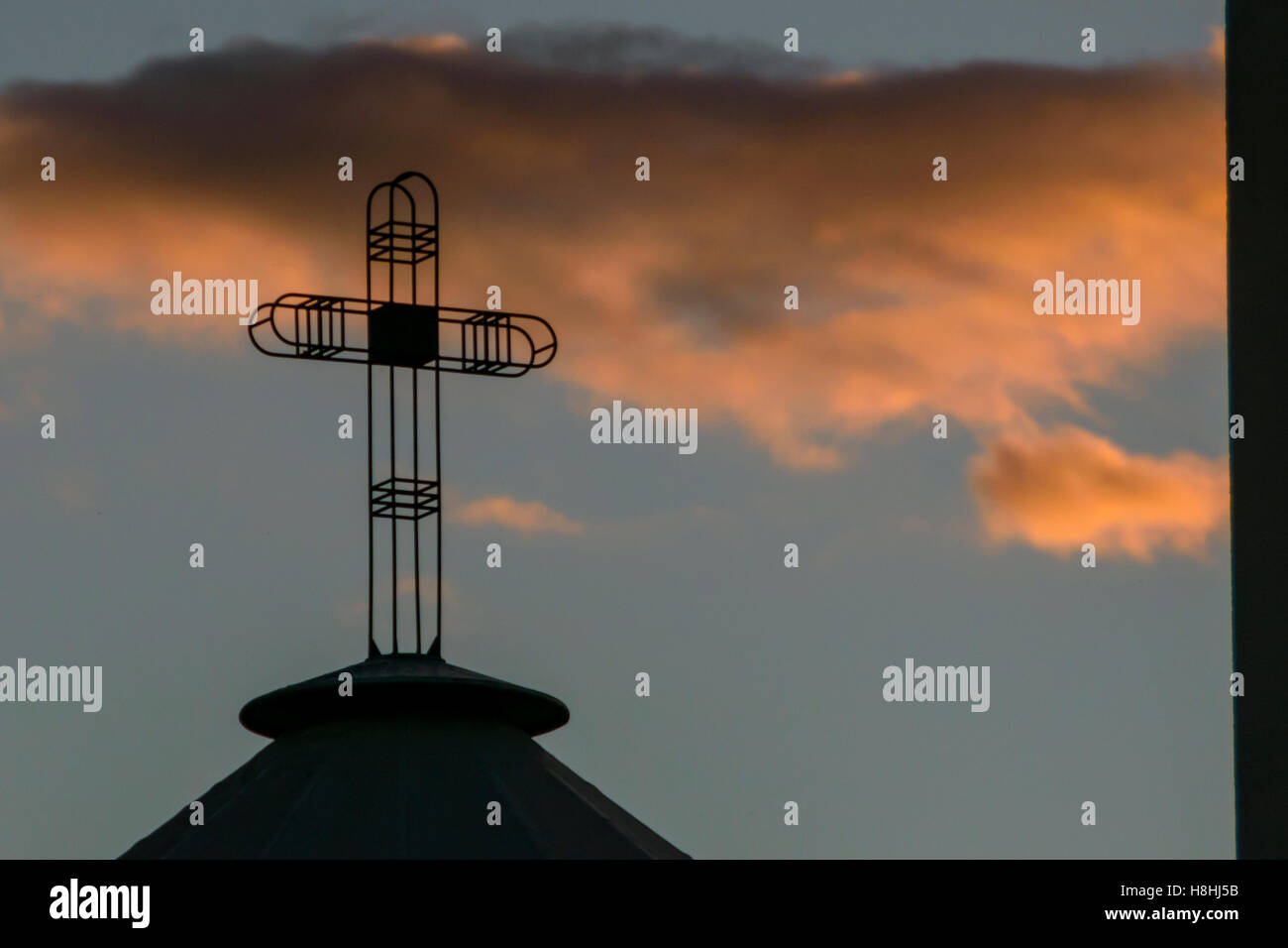 Sihuette eines modernen zeitgenössischen christlichen Kreuzes oben auf der Kirche, dramatische Wolken bei Sonnenuntergang als Hintergrund beleuchtet Stockfoto
