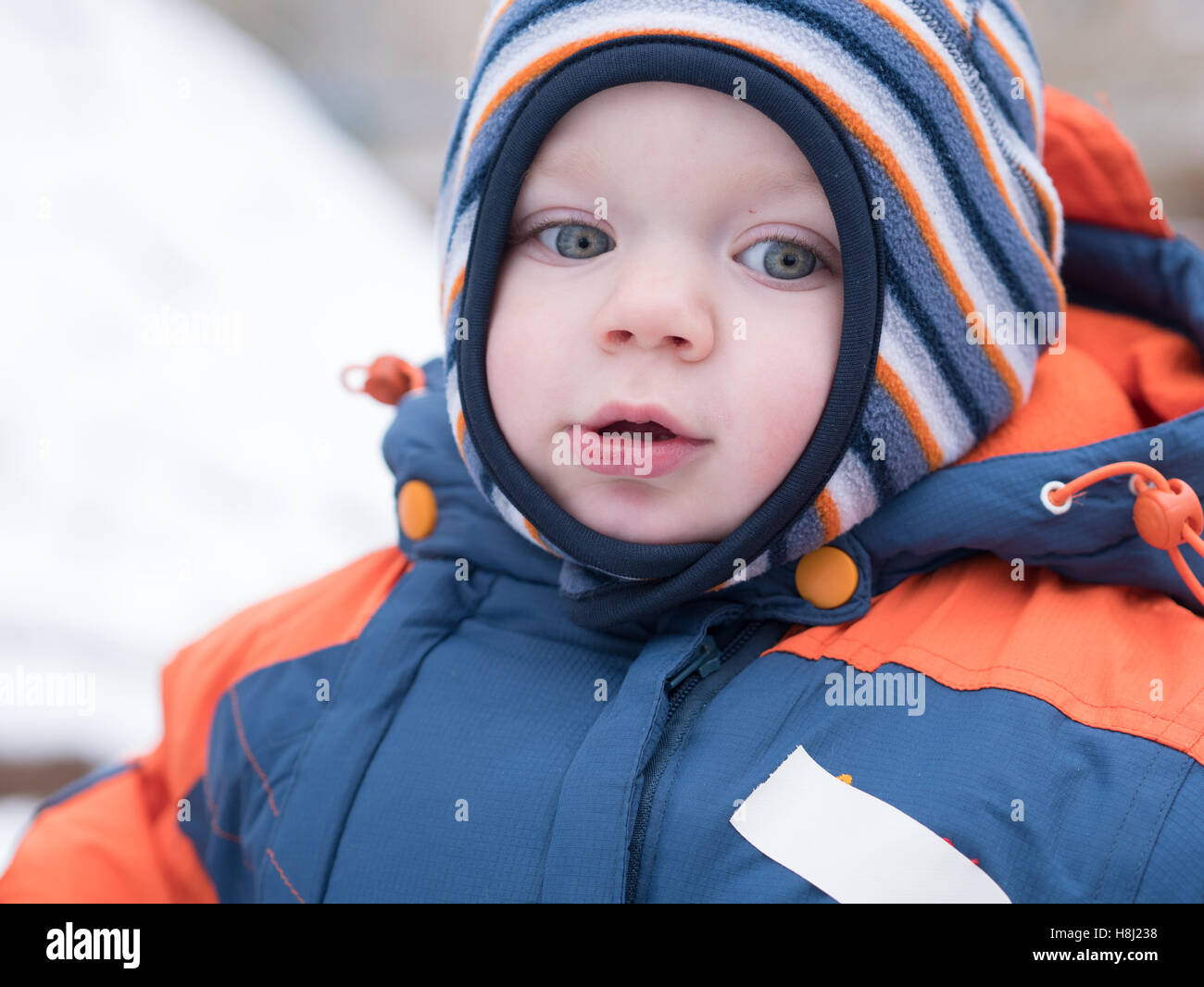 Attraktiven jungen spielen mit dem ersten Schnee. Er lächelt und schaut Schneemann. Dick blau-Orange Overall hell gestreiften Hut auf ein Jahr altes Kind. Stockfoto