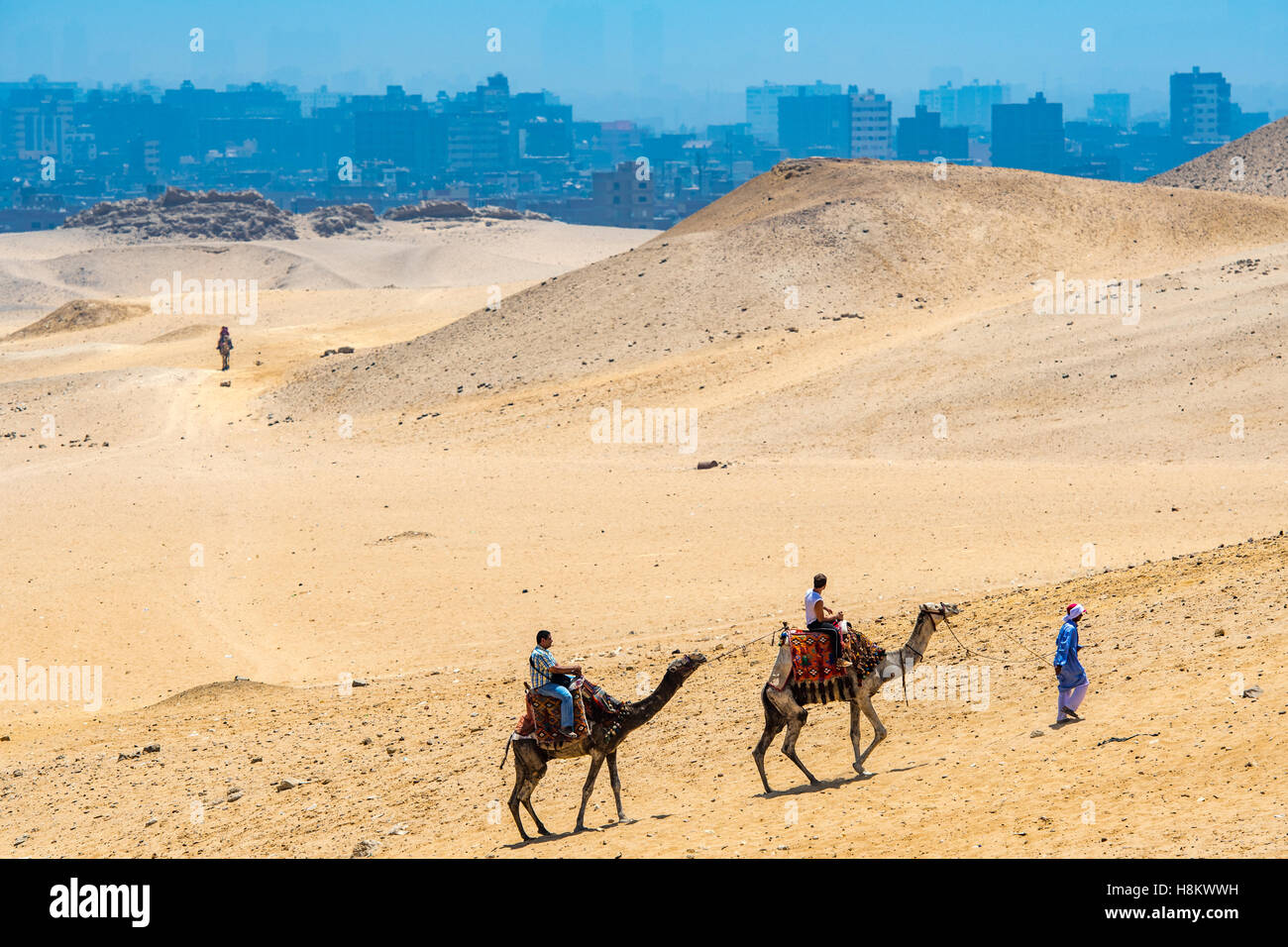 Kairo, Ägypten Kamel Fahrer und Touristen zu Fuß durch die Wüste mit Kairos im Hintergrund Kamele reiten. Stockfoto