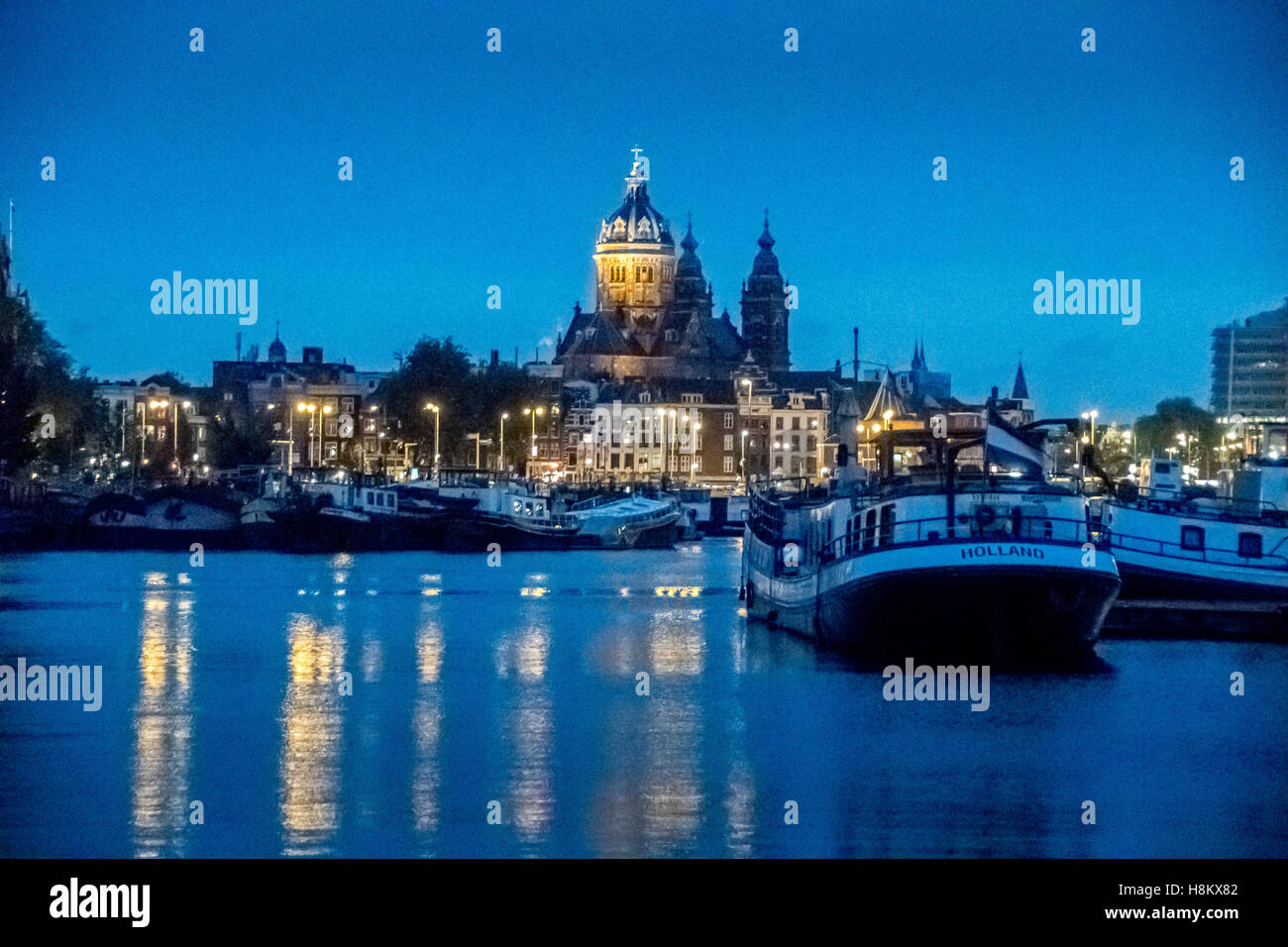 Amsterdam, Niederlande-Boote im Kanal mit Wasser-Ansicht der St. Nikolaus-Kathedrale in der Nacht angedockt. Stockfoto