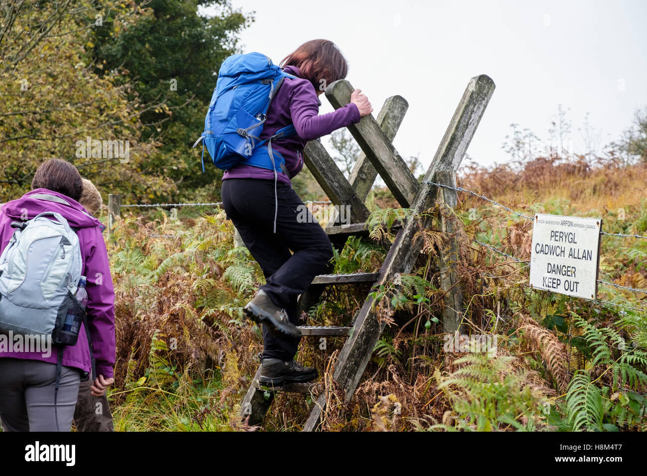 Zweisprachige "Gefahr fernzuhalten" Zeichen auf einem öffentlichen Wanderweg bei Wanderern einen Leiter-Stil über einen Stacheldrahtzaun zu klettern. Wales UK Stockfoto