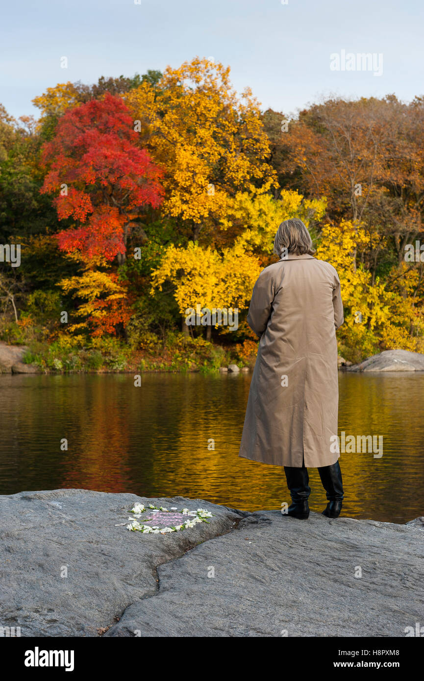 City Park, Central Park, New York City, USA, eine einsame Frau mittleren Alters, die auf Felsen am Ufer eines Sees steht und einen Regenmantel trägt Stockfoto