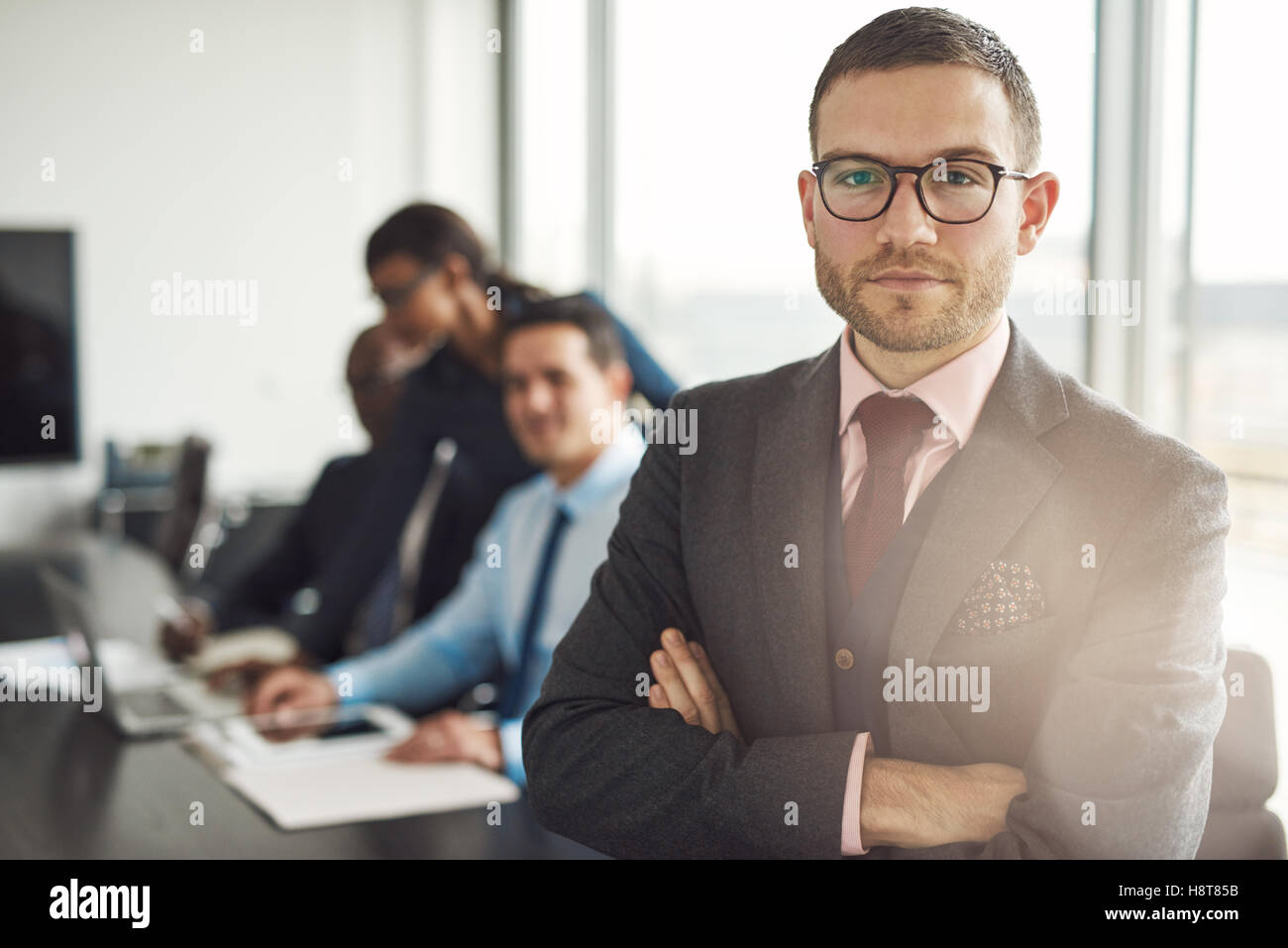 Schöne bärtige Professional in Brillen und Krawatte stehen mit verschränkten Armen neben drei beschäftigten Kollegen vor großen Offi Stockfoto