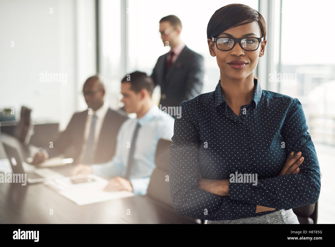 Überzeugt junge afrikanische Geschäftsfrau stehend mit gefalteten Arme lächelnd in die Kamera in einem Sitzungssaal mit männlichen Kollegen in der Stockfoto