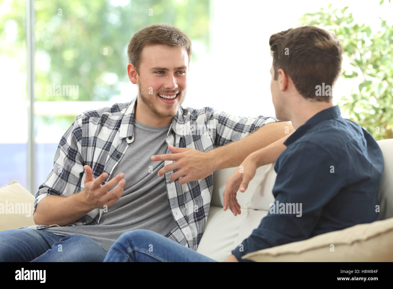 Zwei Freunde sitzen in einer Couch im Wohnzimmer mit einem Fenster im Hintergrund zu Hause sprechen Stockfoto