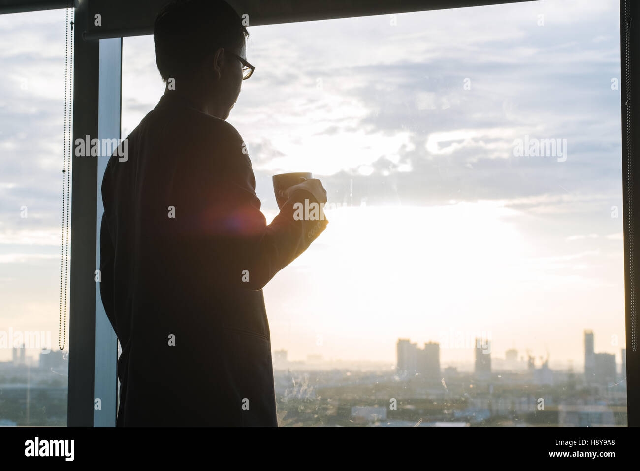 Silhouette eines Geschäftsmannes hält Kaffee und Blick auf die Stadt durch Fenster im Büro Stockfoto