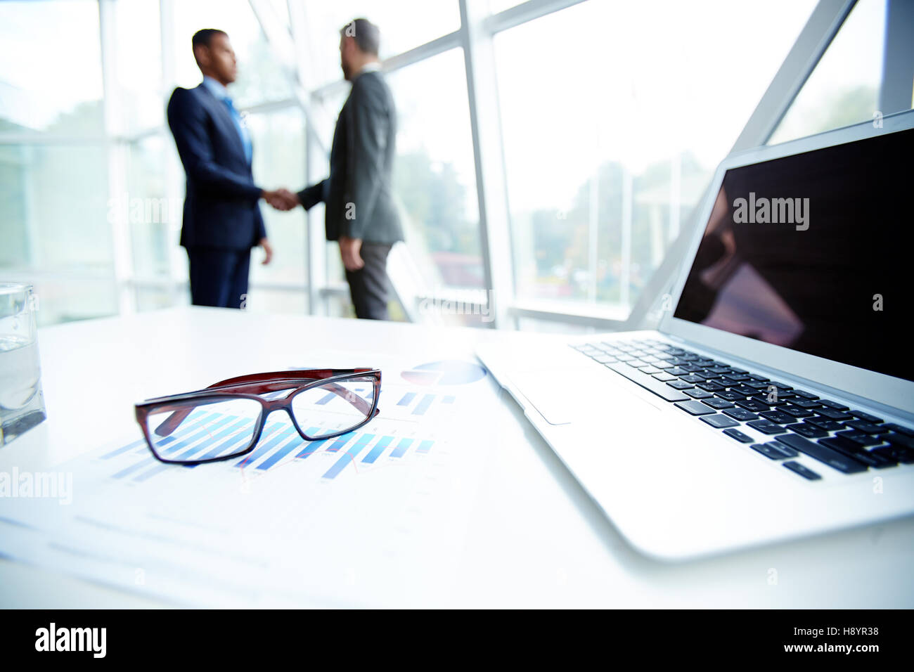 Arbeitsplatz der Finanzmanager mit Brille, Laptop und Dokument auf Grund von zwei Männern handshaking Stockfoto