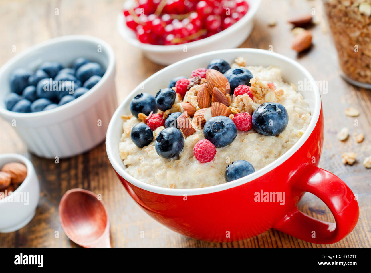 Haferflocken-Porridge mit frischen Heidelbeeren, Himbeeren, Müsli und Mandeln in der roten Schale am weißen Tisch. Gesundes Frühstück Stockfoto