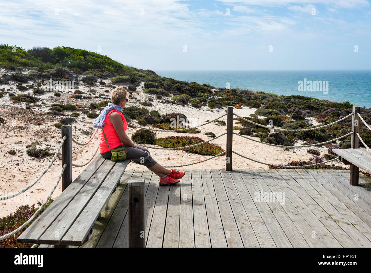 Erwachsene Frau mit blonden Haaren und orangefarbenen t-Shirt sitzt an der Küste von Portugal-Alentejo und Blick über den Horizont Stockfoto