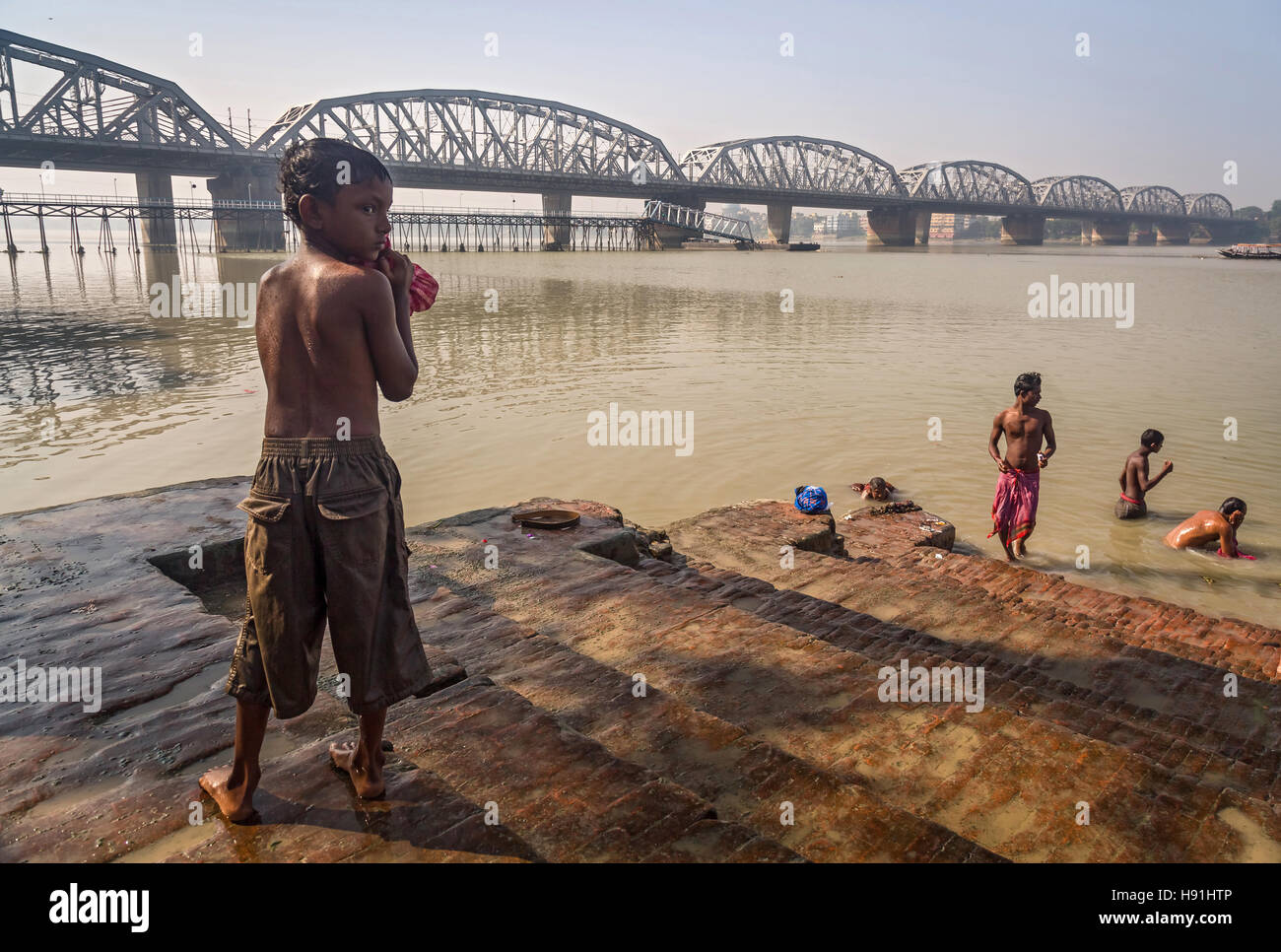 Straßenkind nach einem Bad im Fluss Ganges bei Dakshineshwar Baden Ghat mit der Bally-Brücke vor dem Hintergrund. Stockfoto