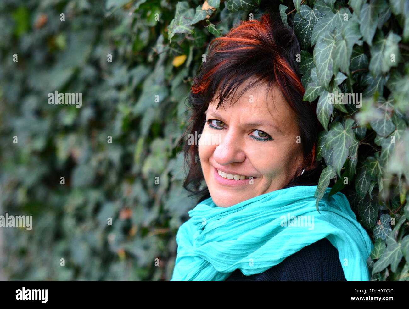 Porträt von Smilling mittleren gealterten gewöhnliche kaukasischen Frau mit langen, dunklen Haaren und schwarzen Pulli an Efeu Wand gelehnt. Stockfoto