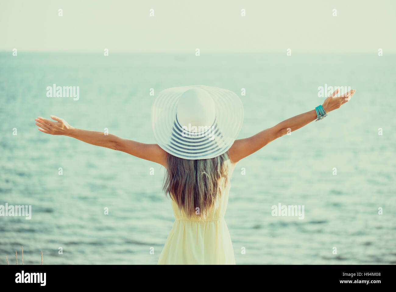 Glückliche junge Frau trägt in Kleid und Hut mit eine breite Krempe mit erhobenen Armen stehend und Aussicht auf Meer, Sicht nach hinten. Getönten Bild Stockfoto