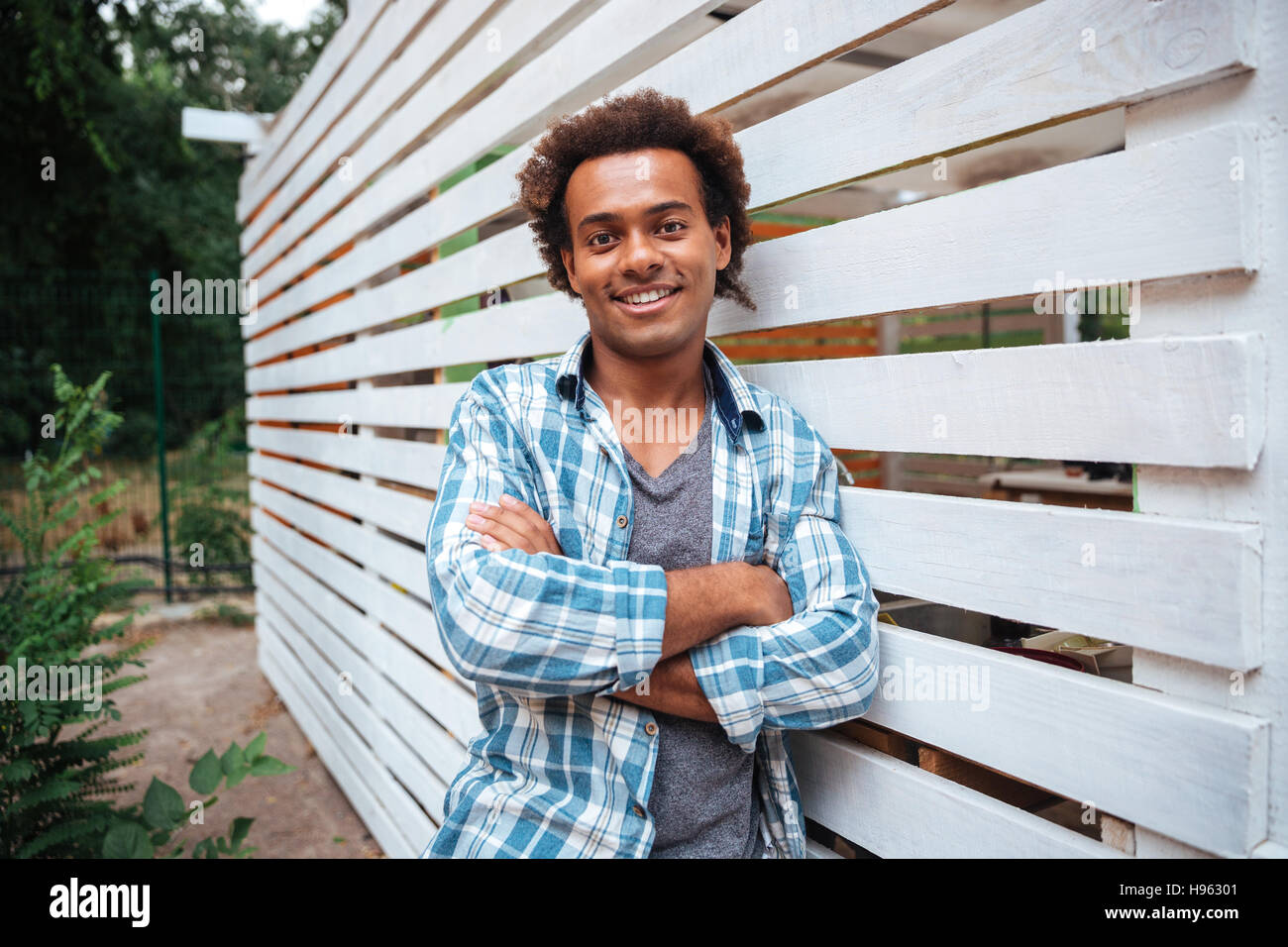 Lächelnd attraktive afrikanische jungen Mann im karierten Hemd stehen im Freien mit Arme gekreuzt Stockfoto
