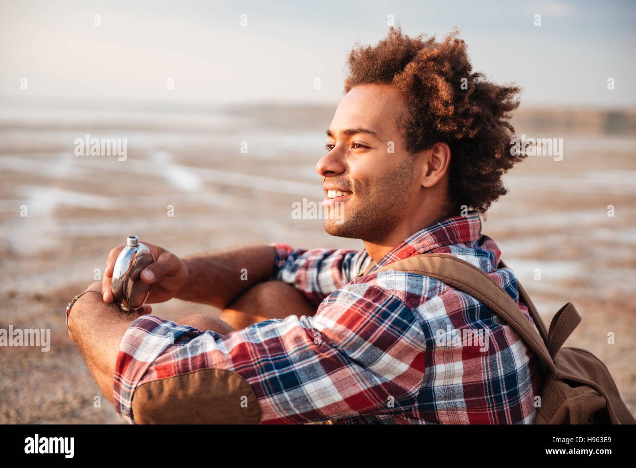 Lächelnd afrikanischen jungen Mann mit Rucksack aus Flachmann am Strand trinken Stockfoto