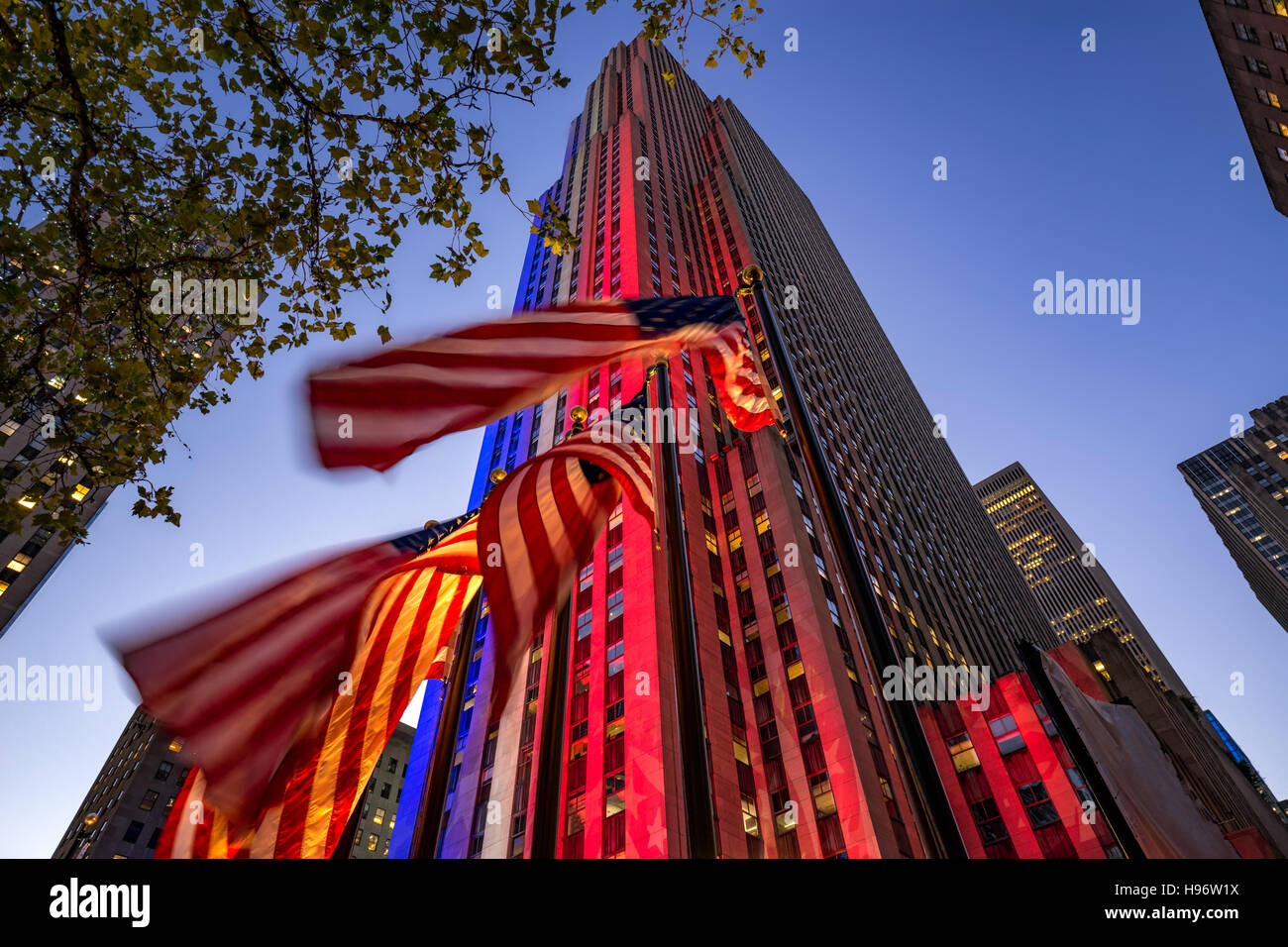 Rockefeller Center in der Dämmerung in weiß, rot und blau beleuchtet. Amerikanische Flaggen flattern im Wind. Midtown Manhattan, New York Stockfoto