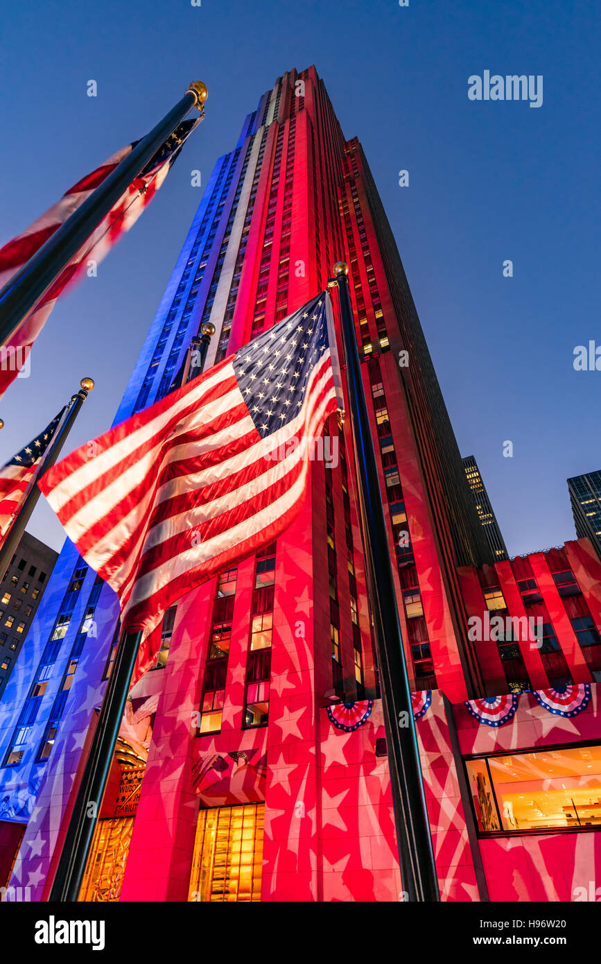 Rockefeller Center in der Dämmerung in weiß, rot und blau beleuchtet. Amerikanische Flaggen flattern im Wind. Midtown Manhattan, New York Stockfoto
