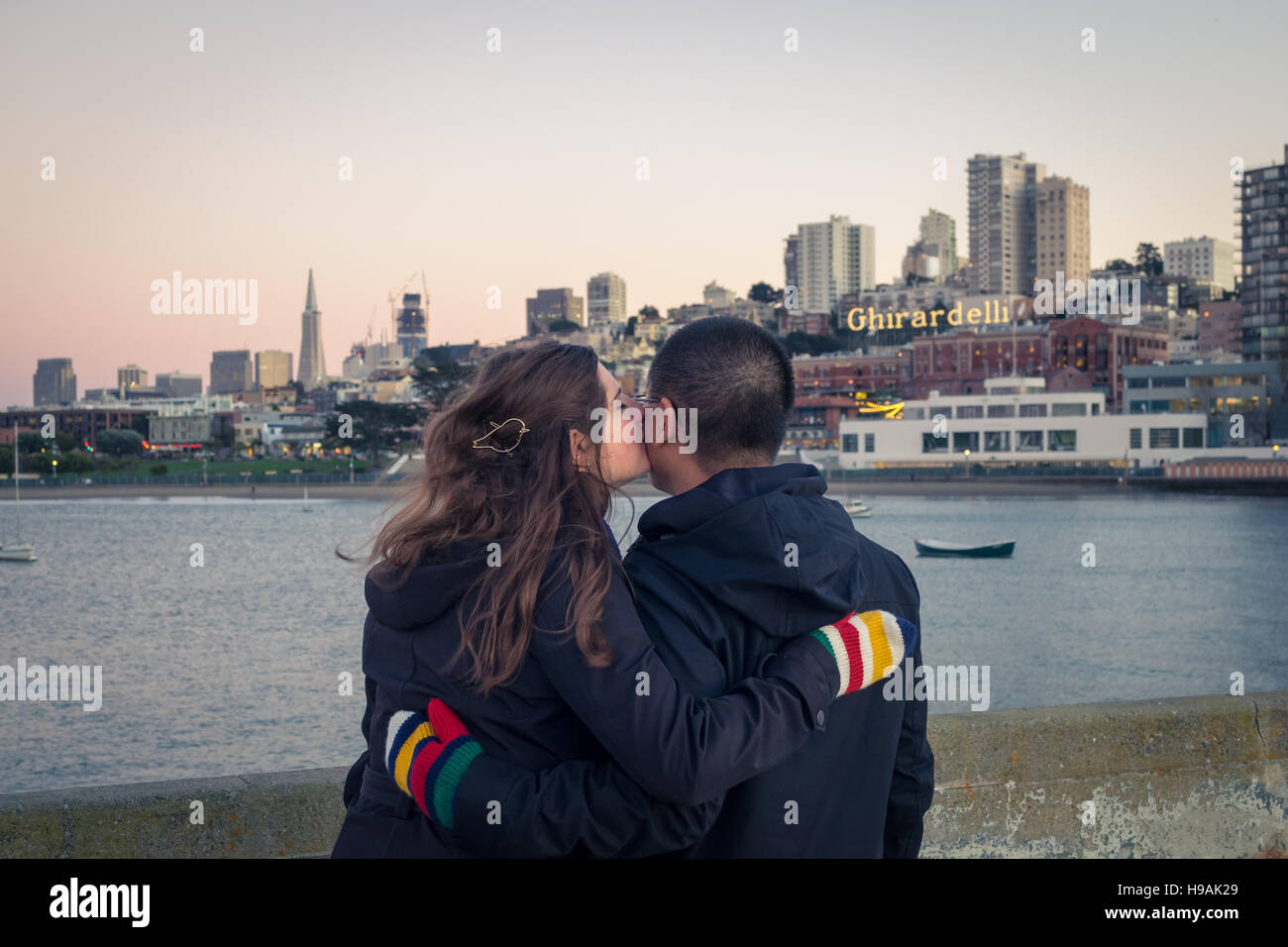 Ein junges Paar in Liebe am Aquatic Park Pier mit der Skyline von Aquatic Park Badehaus, Ghirardelli Square und San Francisco. Stockfoto