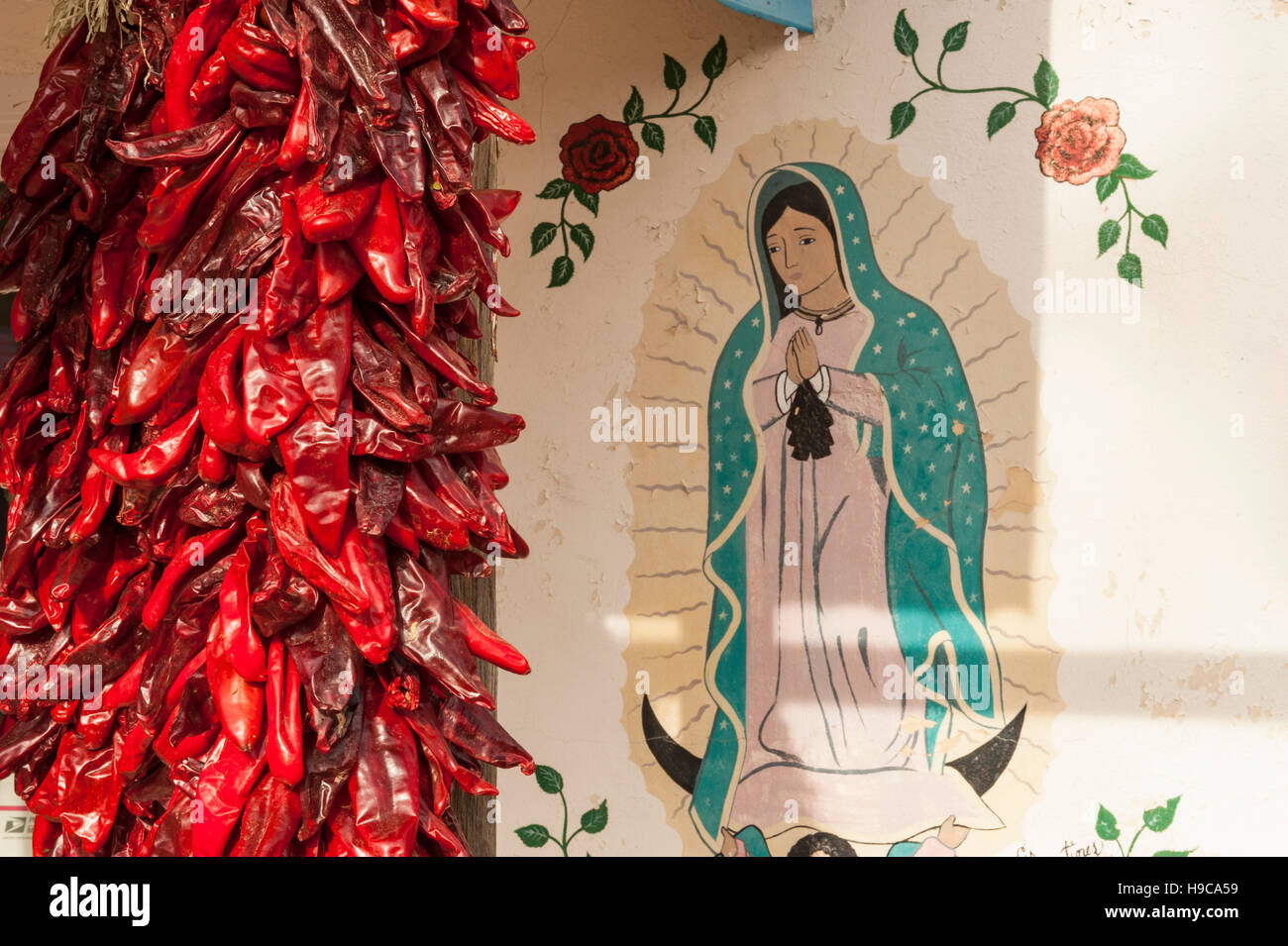 Ein paar rote Chilis (Capsicum Annuum) hängen neben ein Bild der Jungfrau Maria in Chimayo, New Mexico, NM, USA. Stockfoto