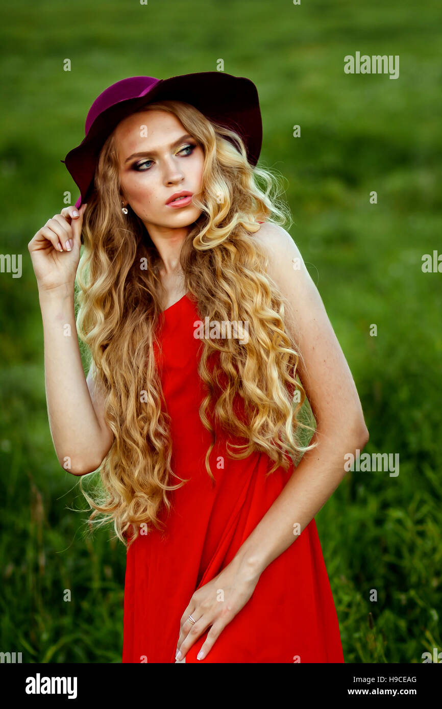 junge schöne Mädchen in einem roten Hut und roten Kleid stehen im Feld auf grünem Grund, blicken nicht in die Kamera Stockfoto