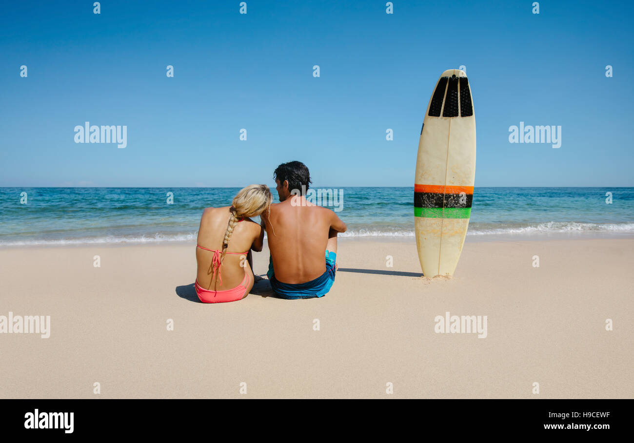 Rückansicht Schuss der junge Mann und Frau sitzen am Strand mit Surfbrett. Paar erholsame am Ufer Meeres an einem Sommertag. Stockfoto