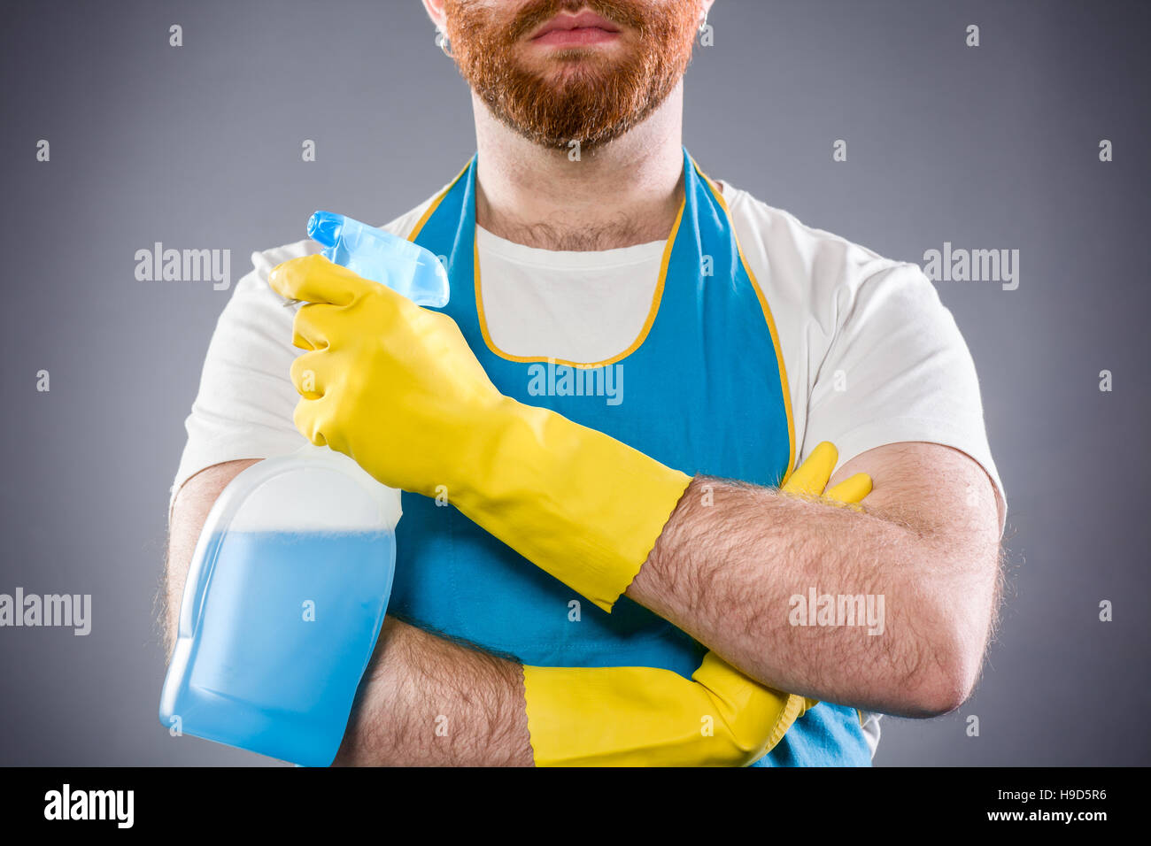 Sauberer Mann mit verschränkten Armen hält ein Waschmittel, eine Schürze und Plastikhandschuhe tragen Stockfoto