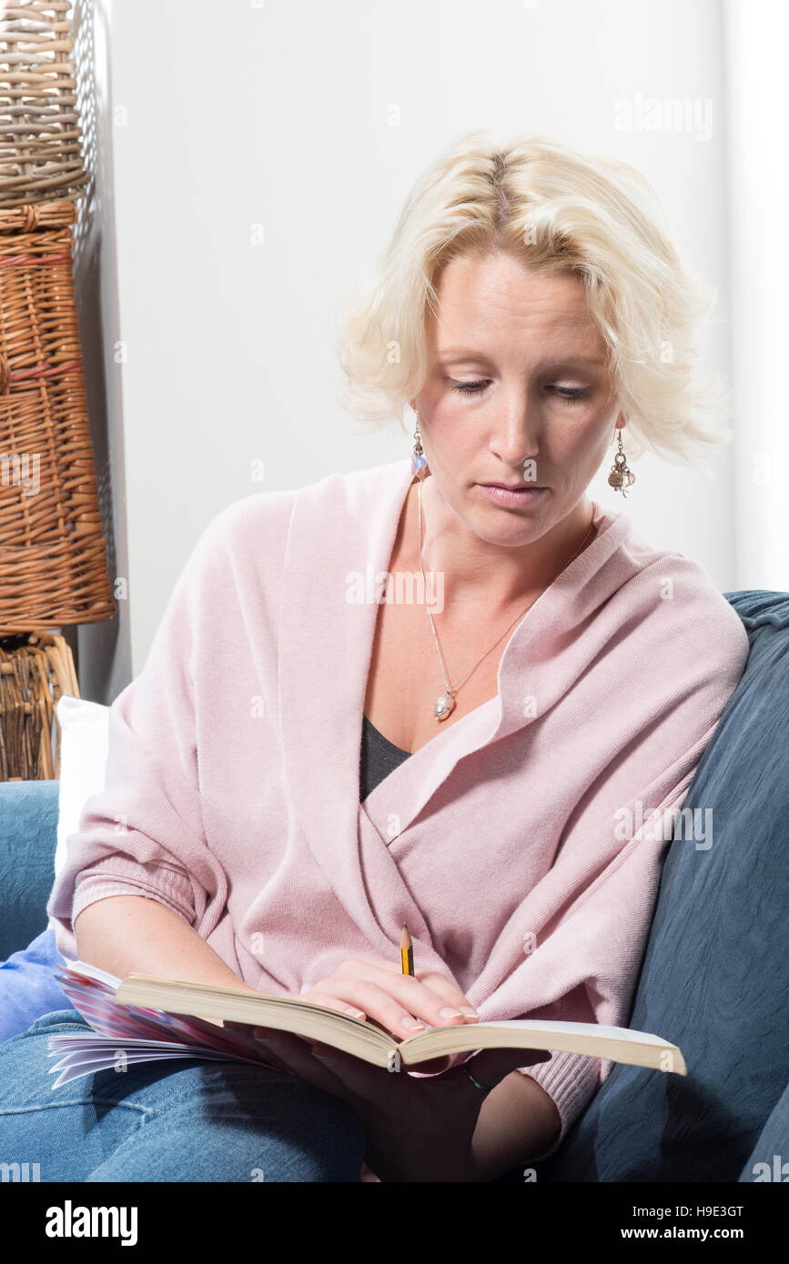 Eine leger gekleidete mittleren gealterte blonde Frau sitzt auf einem Sofa oder Couch hält ein Buch und einen Bleistift. Sie schaut in der Konzentration, wie sie liest. KOPIE-SP Stockfoto