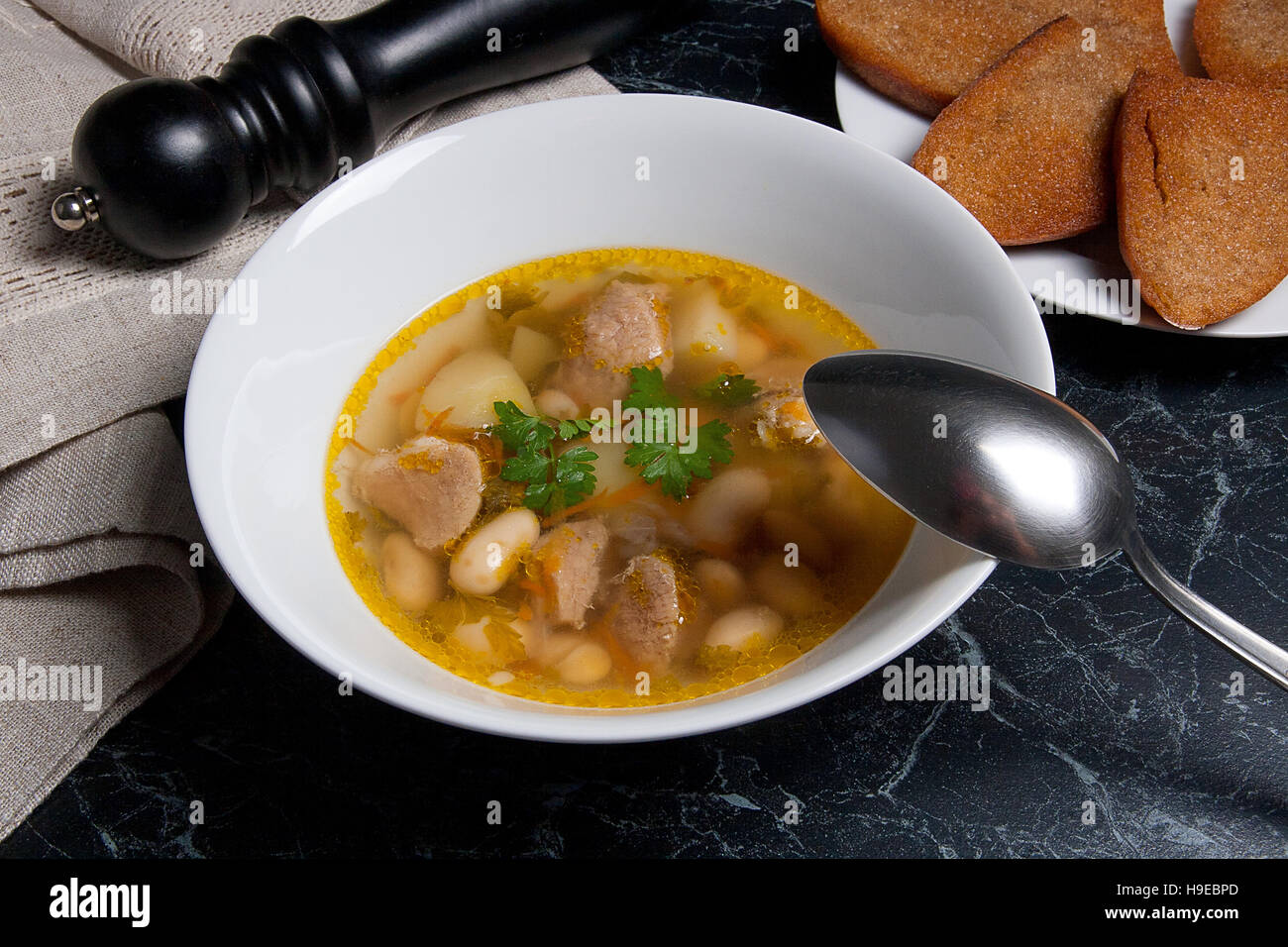 Bohnensuppe in weiße Schüssel mit Metalllöffel auf einem schwarzen Stein Hintergrund. Einige toast auf weißen Teller und schwarze Mühle für Pfeffer auf braunem Tuch. Stockfoto