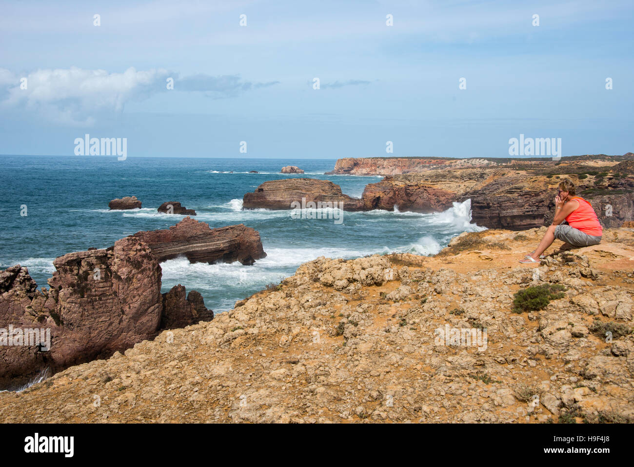 Erwachsenen blonden Frau in orange Shirt sitzen und mit Blick auf die Wellen an der Küste von Portugal in Alentejo Stockfoto