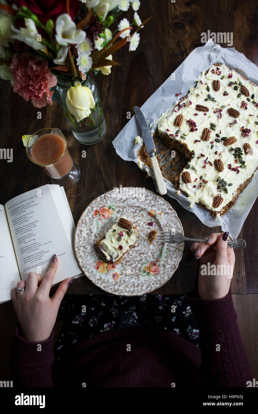 Frau einen Kuchen zu essen, auf dekorative Platte dekoriert mit Pekannüssen & Rosenblätter. Lesung Poesie neben frischen Blumen. Stockfoto