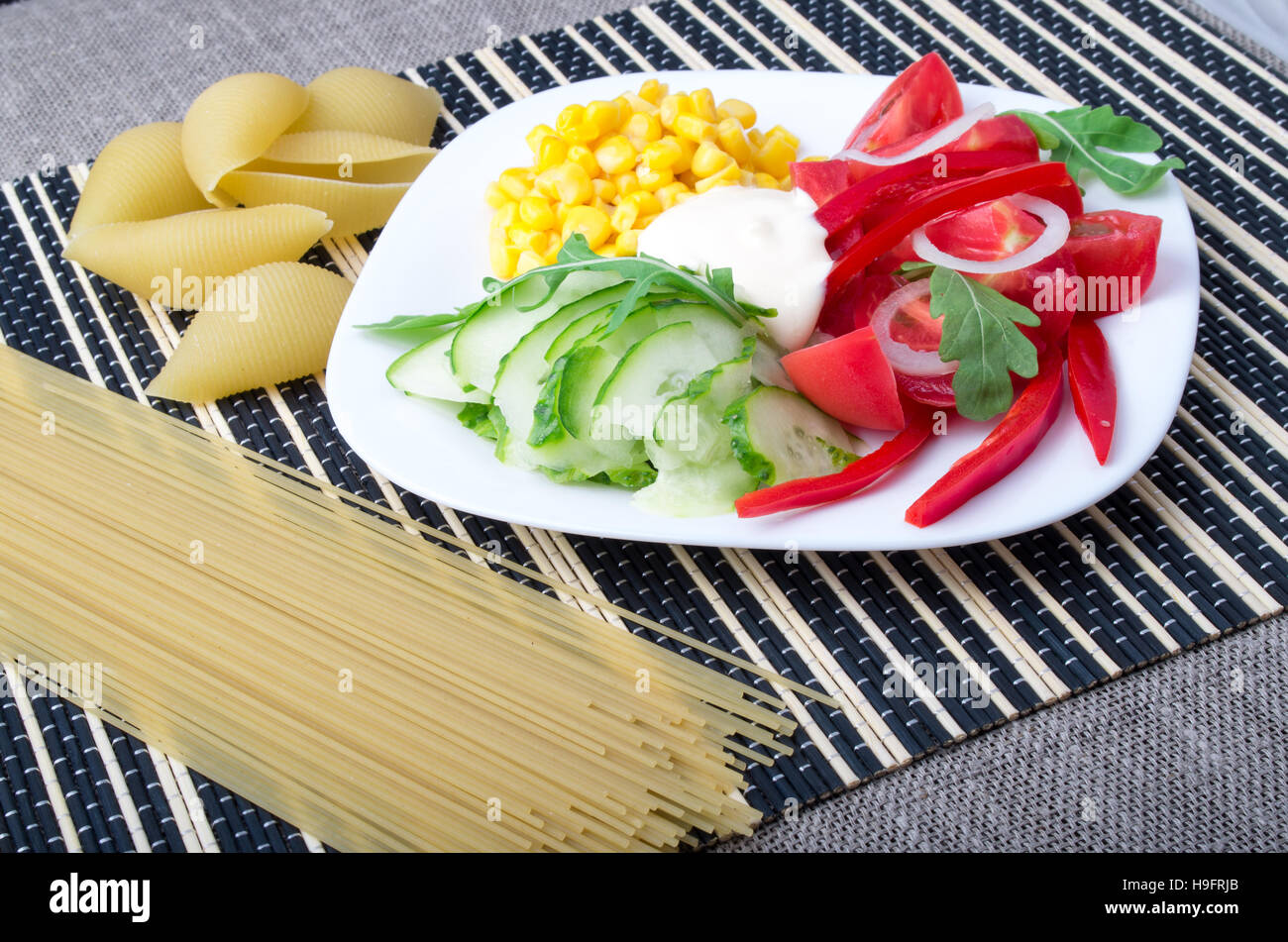 Stücke von frischem rohem Gemüse auf einem weißen Teller - Pfeffer, Tomaten, Mais, Rucola, Gurken, Mozzarella-Käse auf einem gestreiften Hintergrund mit Pasta und sp Stockfoto