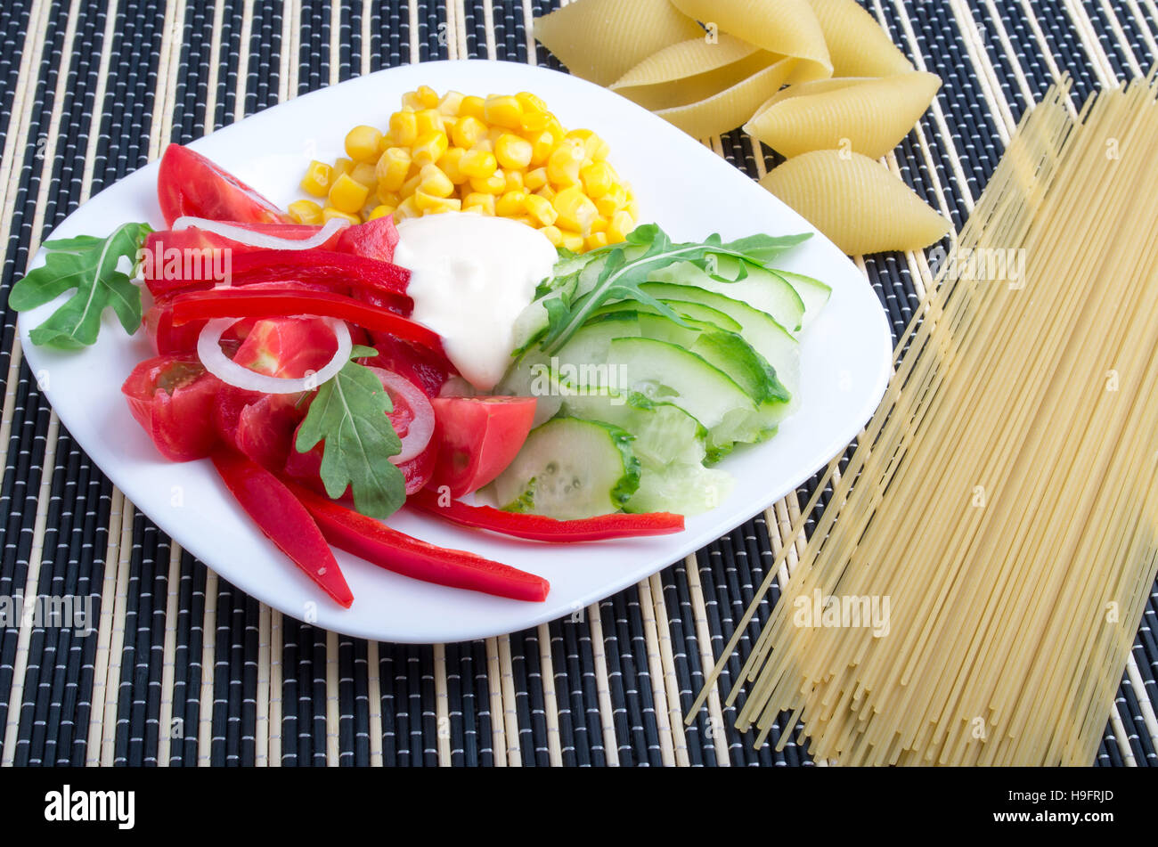 Scheiben von frischem rohem Gemüse auf einem weißen Teller - Pfeffer, Tomaten, Mais, Rucola, Gurken, Mozzarella-Käse auf einem gestreiften Hintergrund mit Pasta und sp Stockfoto