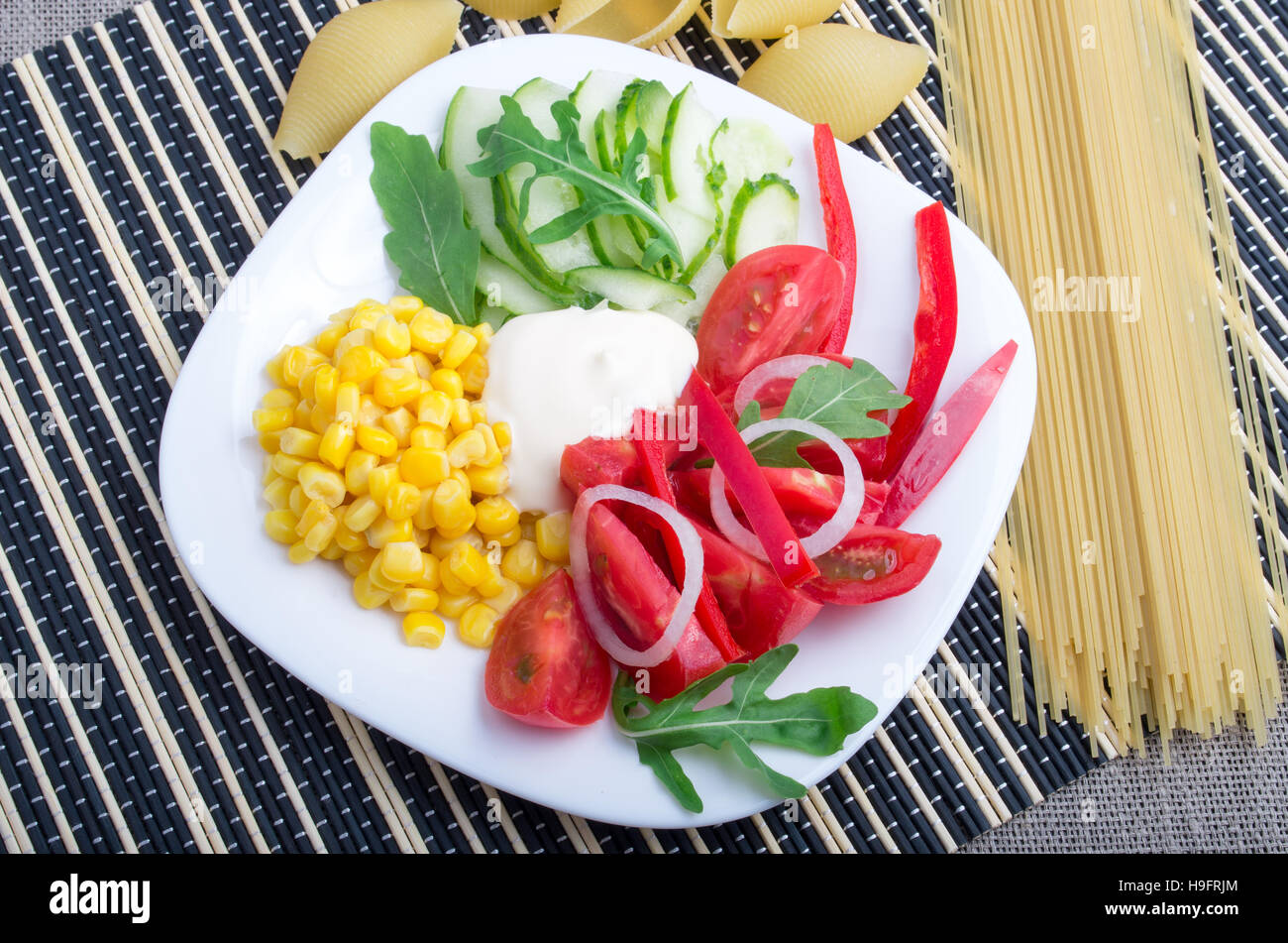 Weißen Teller mit frischen Tomaten, Gurkenscheiben, Mais, Parmeggiano und Zwiebeln neben Zutaten für Spaghetti auf einem gestreiften Hintergrund Stockfoto