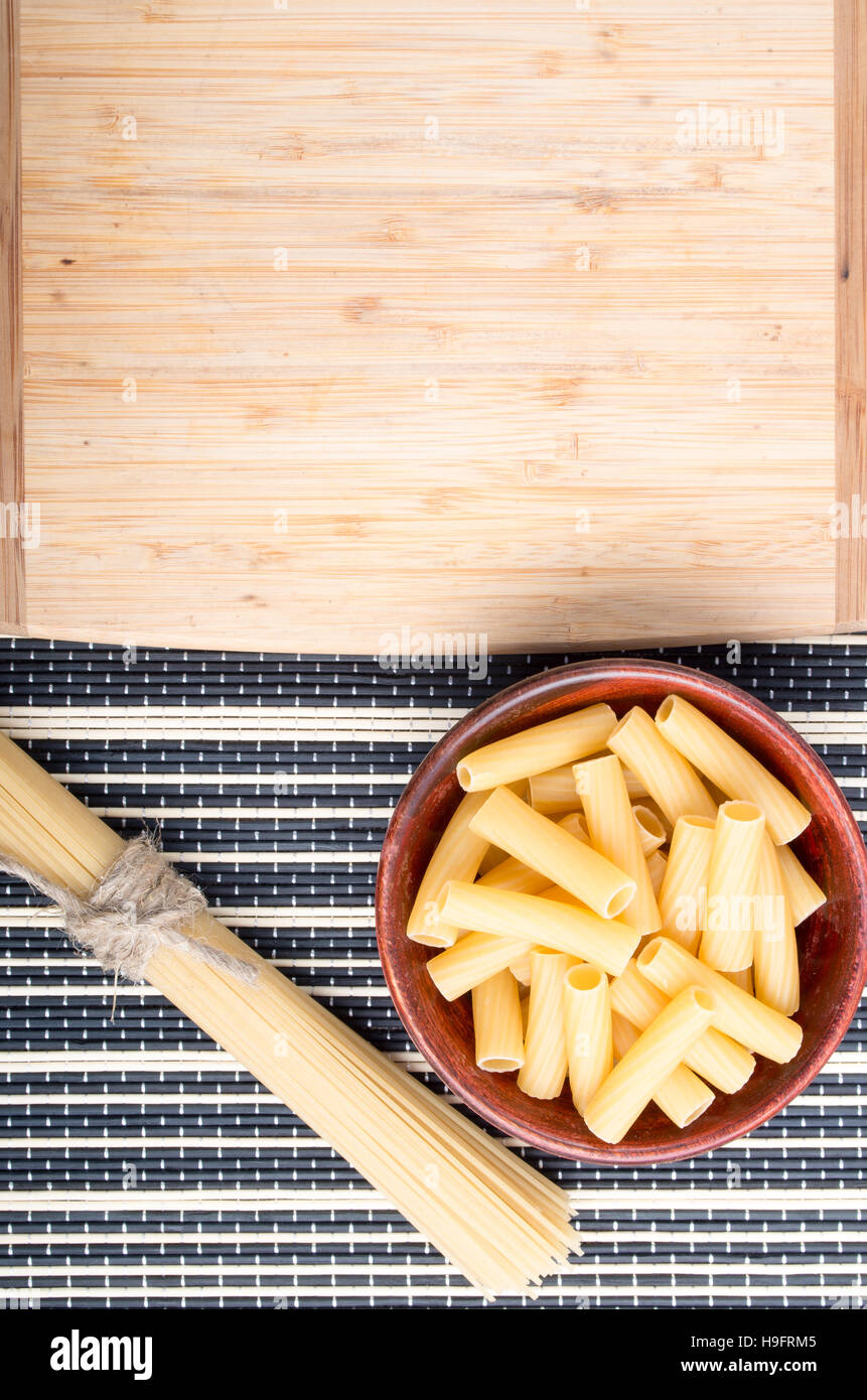 Draufsicht der Nudeln in einer Holzschale mit einem Bündel Spaghetti neben einem Holzbrett zum Kochen auf einem gestreiften Hintergrund Stockfoto